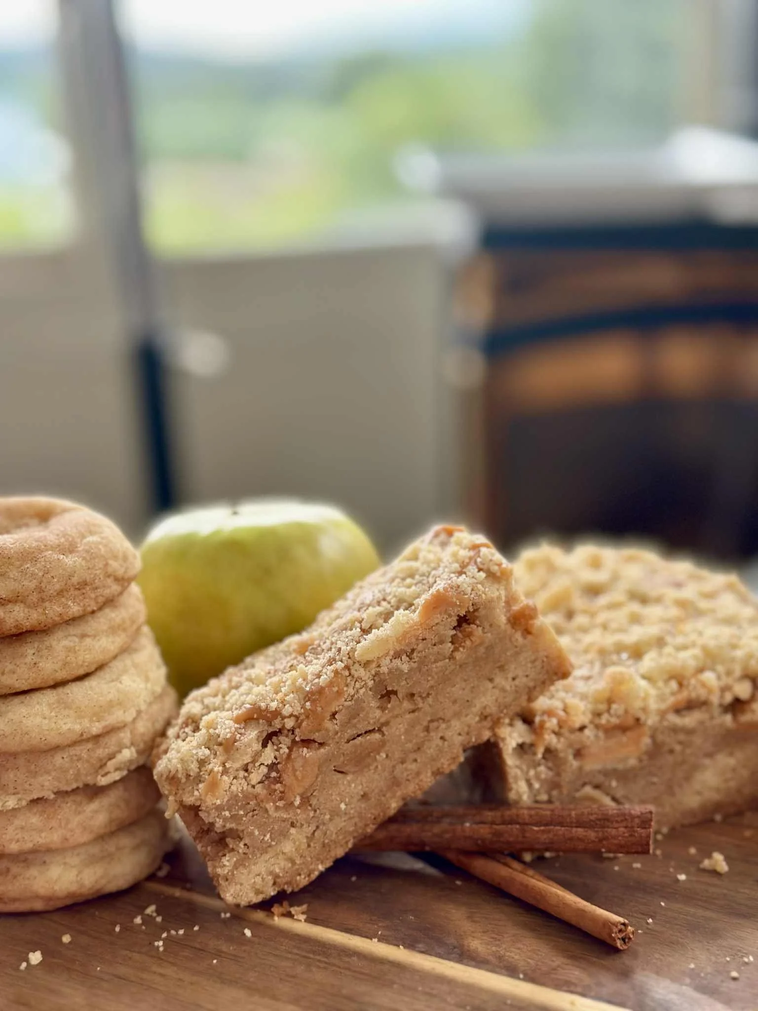 Stack of cookies, a green apple, and a granola bar with cinnamon sticks on a wooden surface near a window