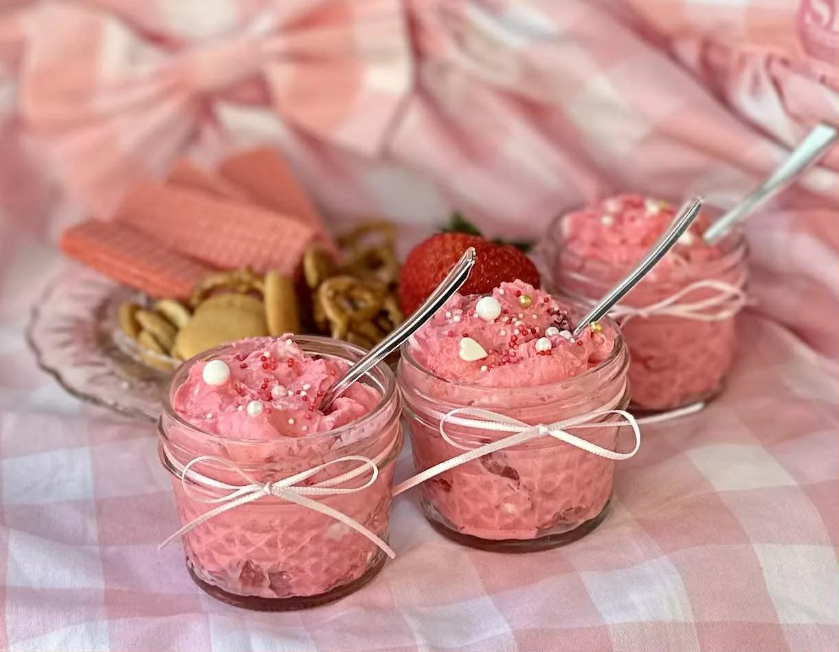 Three glass jars filled with pink strawberry or raspberry-flavored mousse, topped with sprinkles, pearls, and heart-shaped decorations, placed on a pink checkered tablecloth with strawberries and cookies in the background.