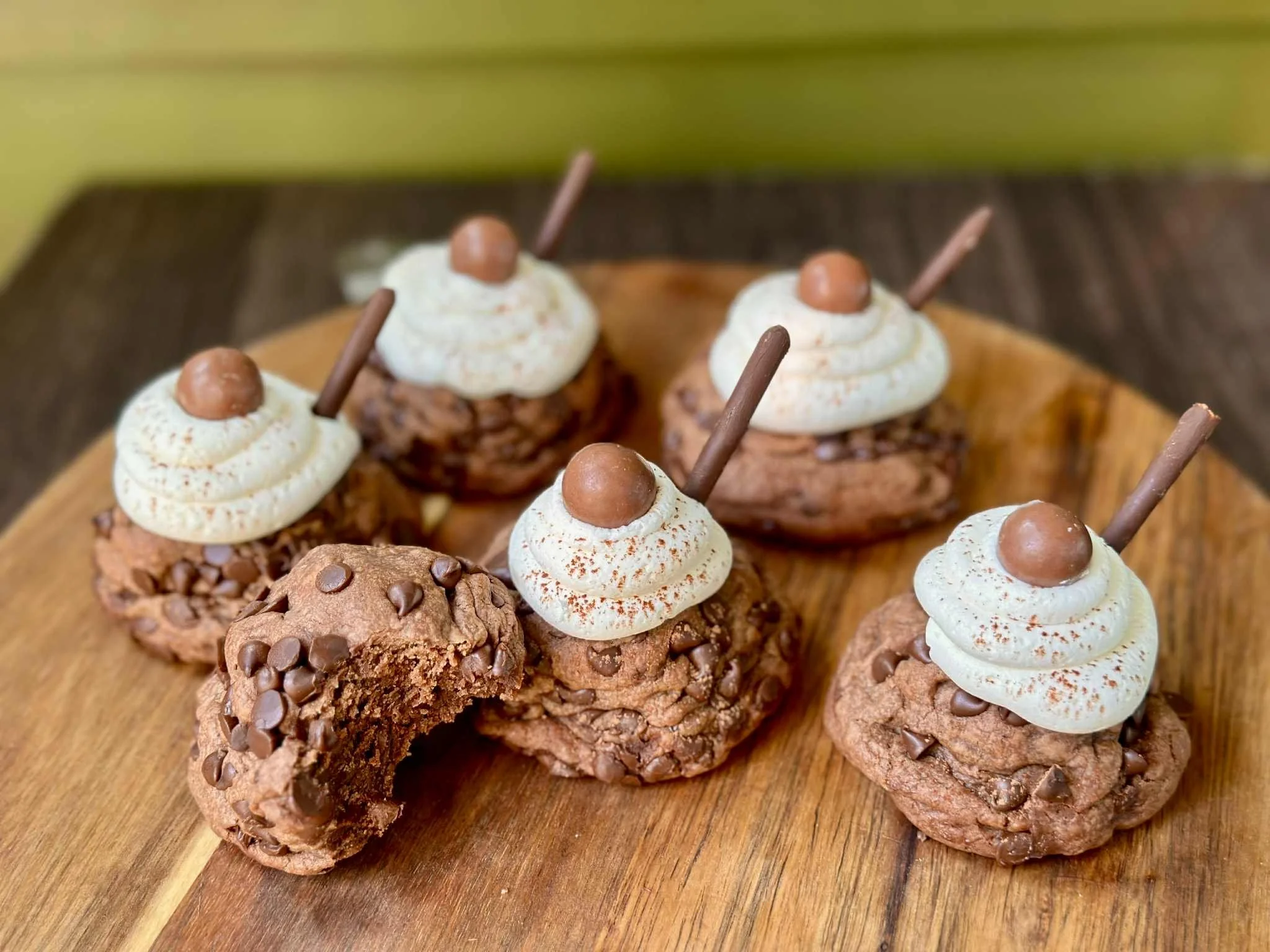 Chocolate cookies topped with whipped cream, a chocolate ball, and a chocolate stick, arranged on a wooden plate.