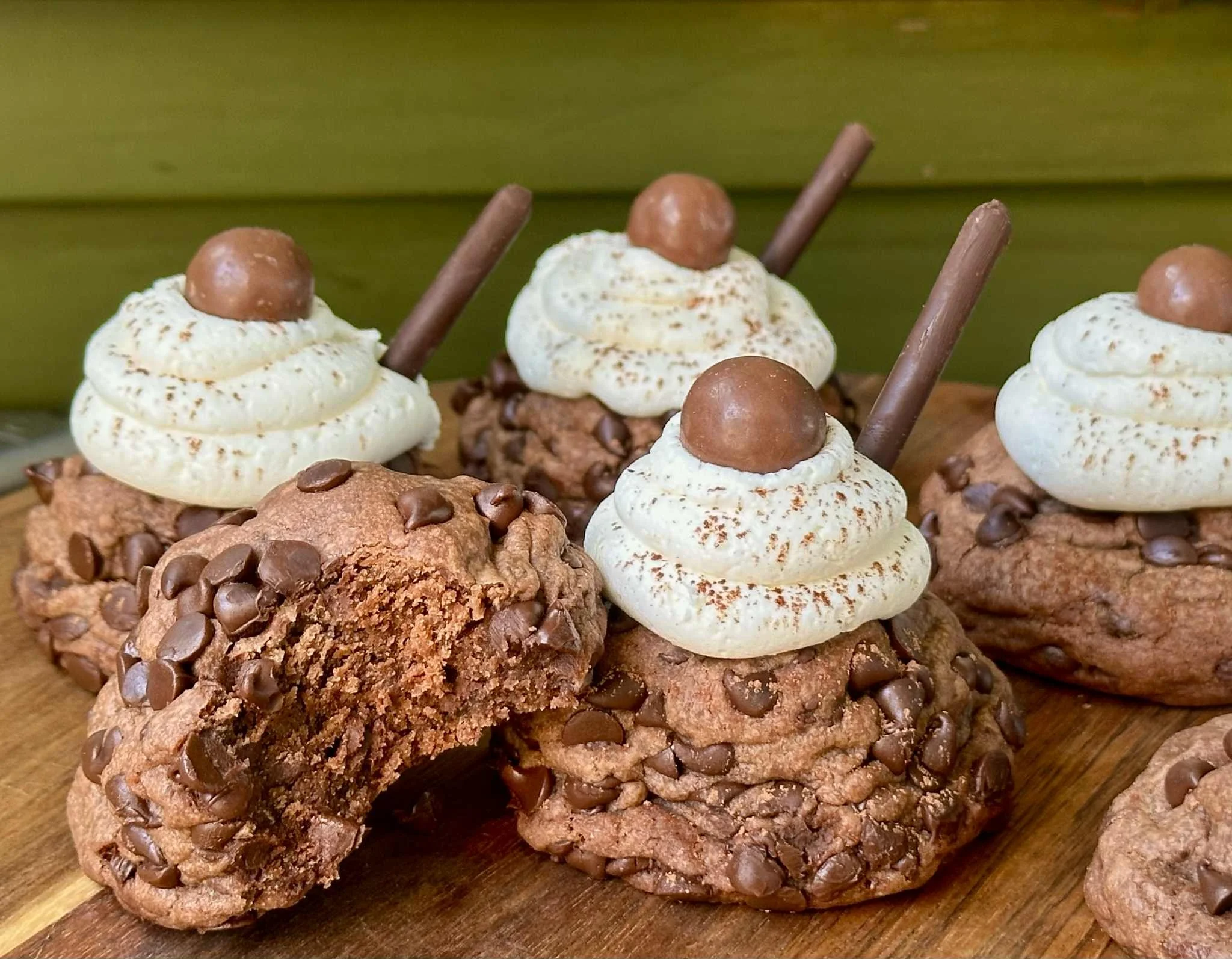 Chocolate chip cookies topped with whipped cream, chocolates, and a chocolate stick, with one cookie bitten into showing the inside.