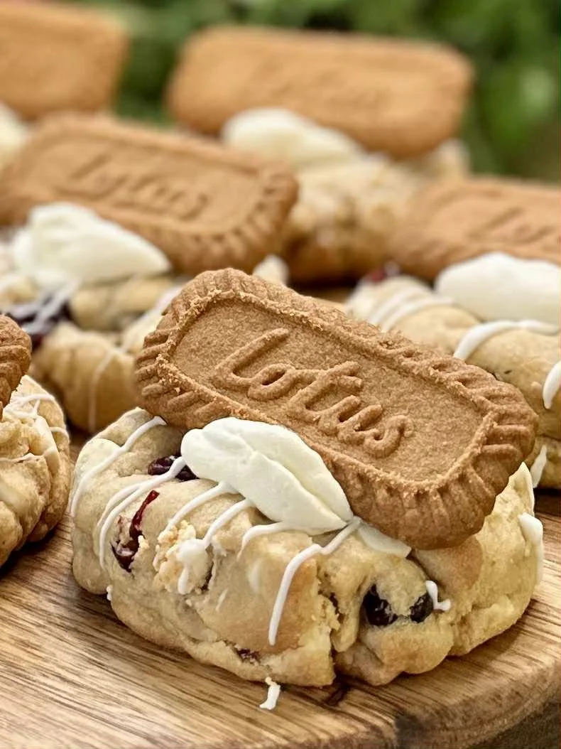 Close-up of a cookie sandwich with cream filling and a biscuit on top labeled 'Lzus', placed on a wooden surface with more cookie sandwiches in the background.