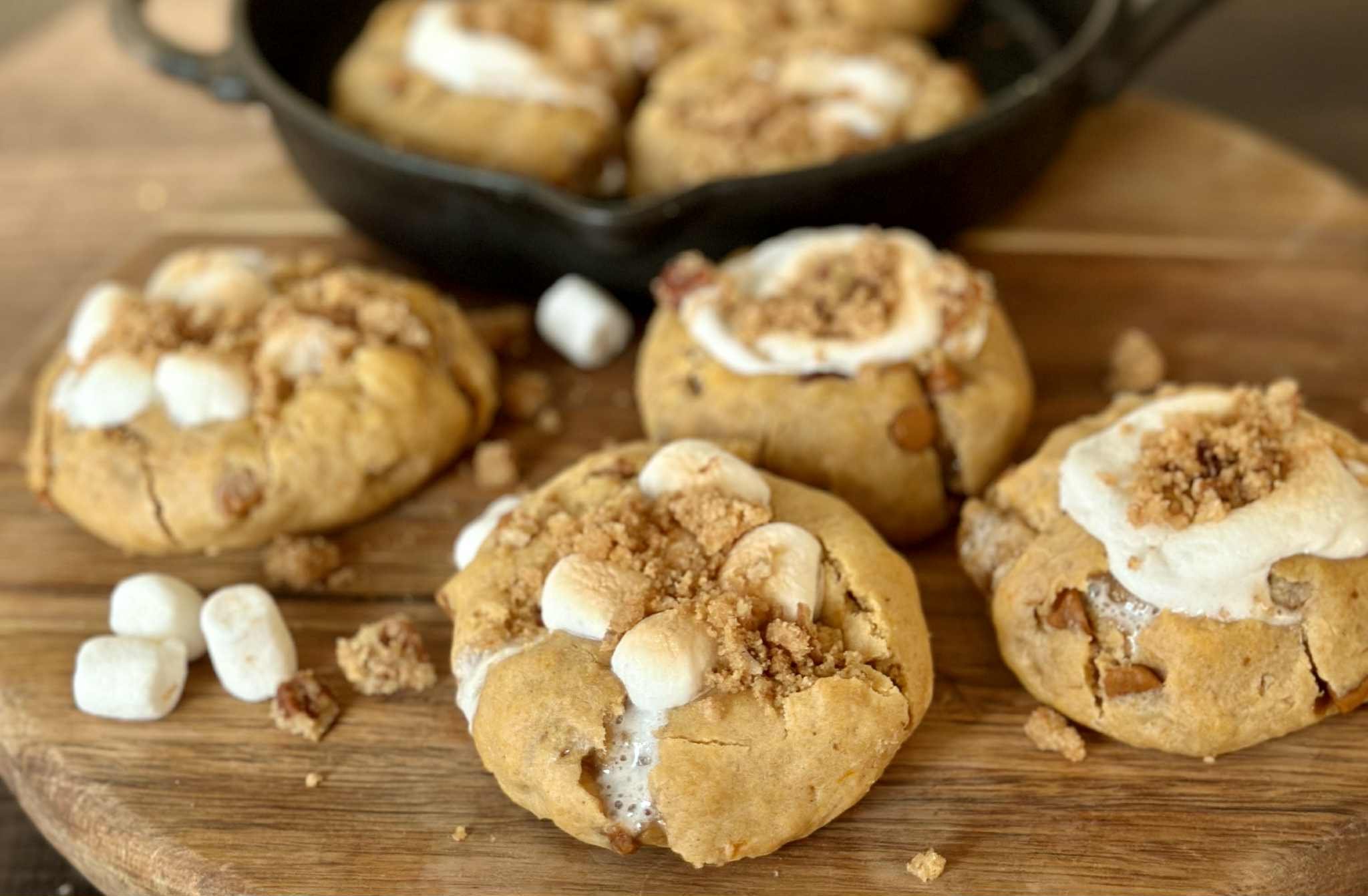 Close-up of four s'mores cookies with marshmallows, graham cracker crumbs, and chocolate, on a wooden board with a black skillet in the background.