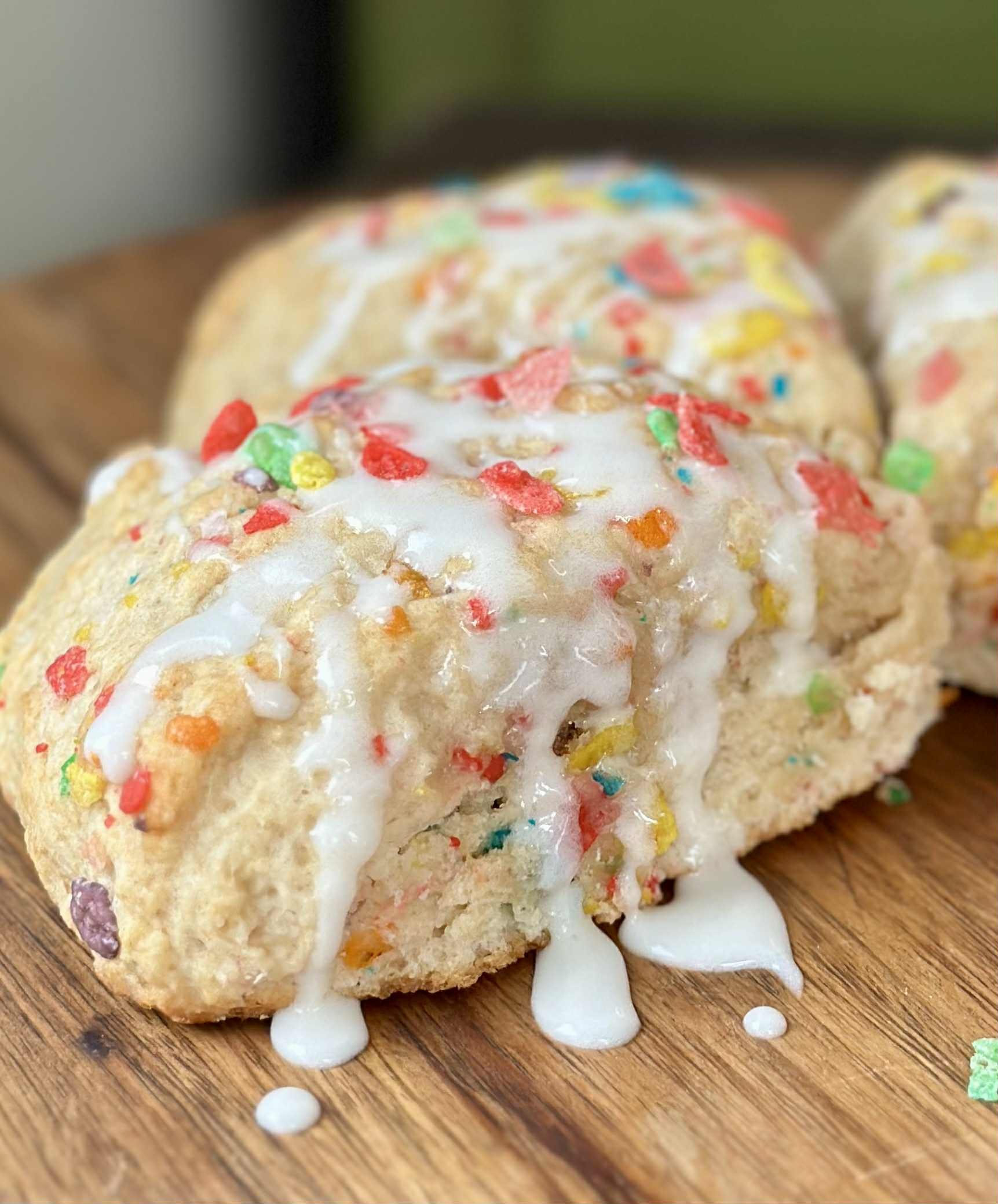 A close-up of a colorful cookie with white icing and rainbow sprinkles, partially broken on a wooden surface.