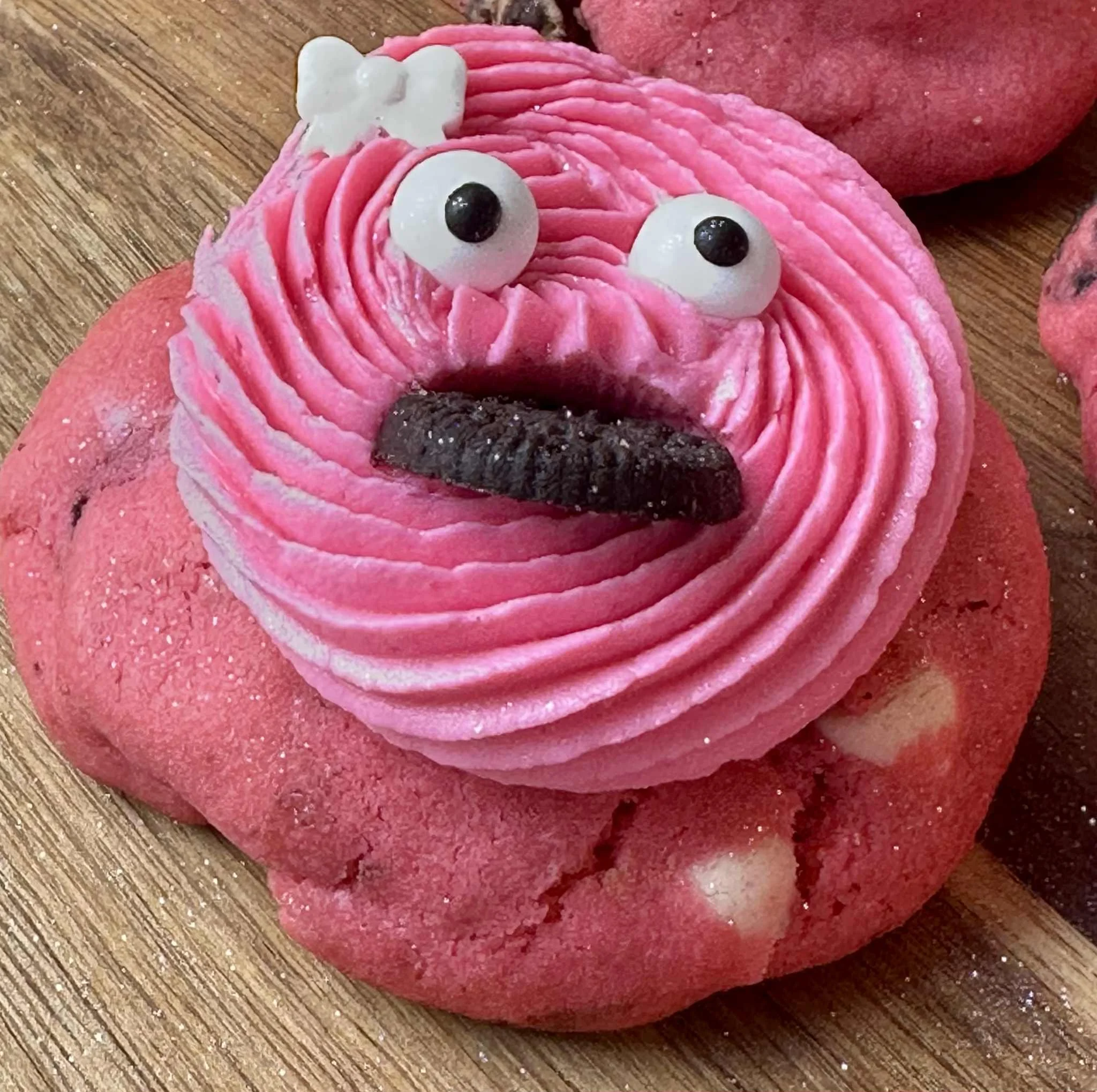 Decorative pink cookie decorated to resemble a face, with piped pink icing, googly eyes, and a small chocolate cookie for a tongue, on a wooden surface.