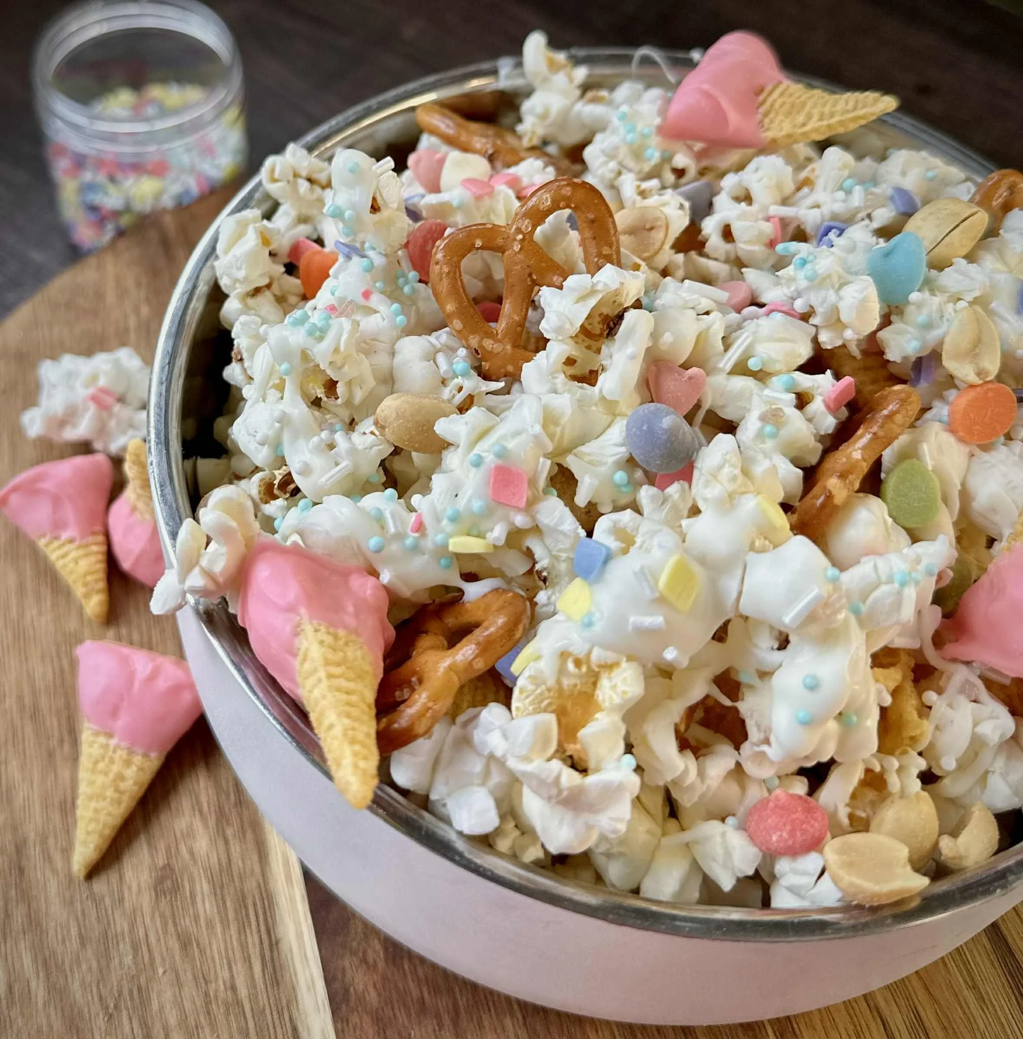 A bowl filled with popcorn decorated with colorful sprinkles, candy hearts, mini ice cream cone-shaped candies with pink icing, pretzels, and scattered candy and baking decorations.