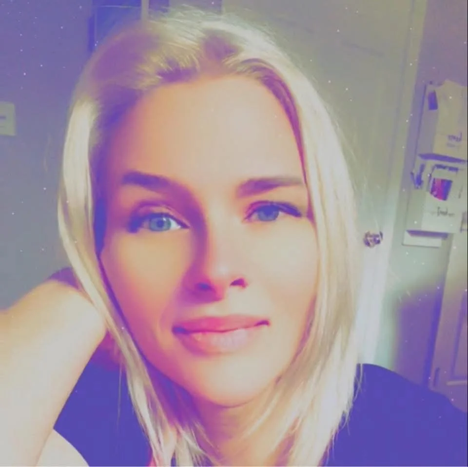 Close-up selfie of a woman with blonde hair and blue eyes, smiling slightly, with a background that appears to be a room with a gray door and electrical panel.