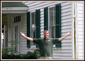 Chuck Moore standing in front of a white house with green shutters, arms spread wide.