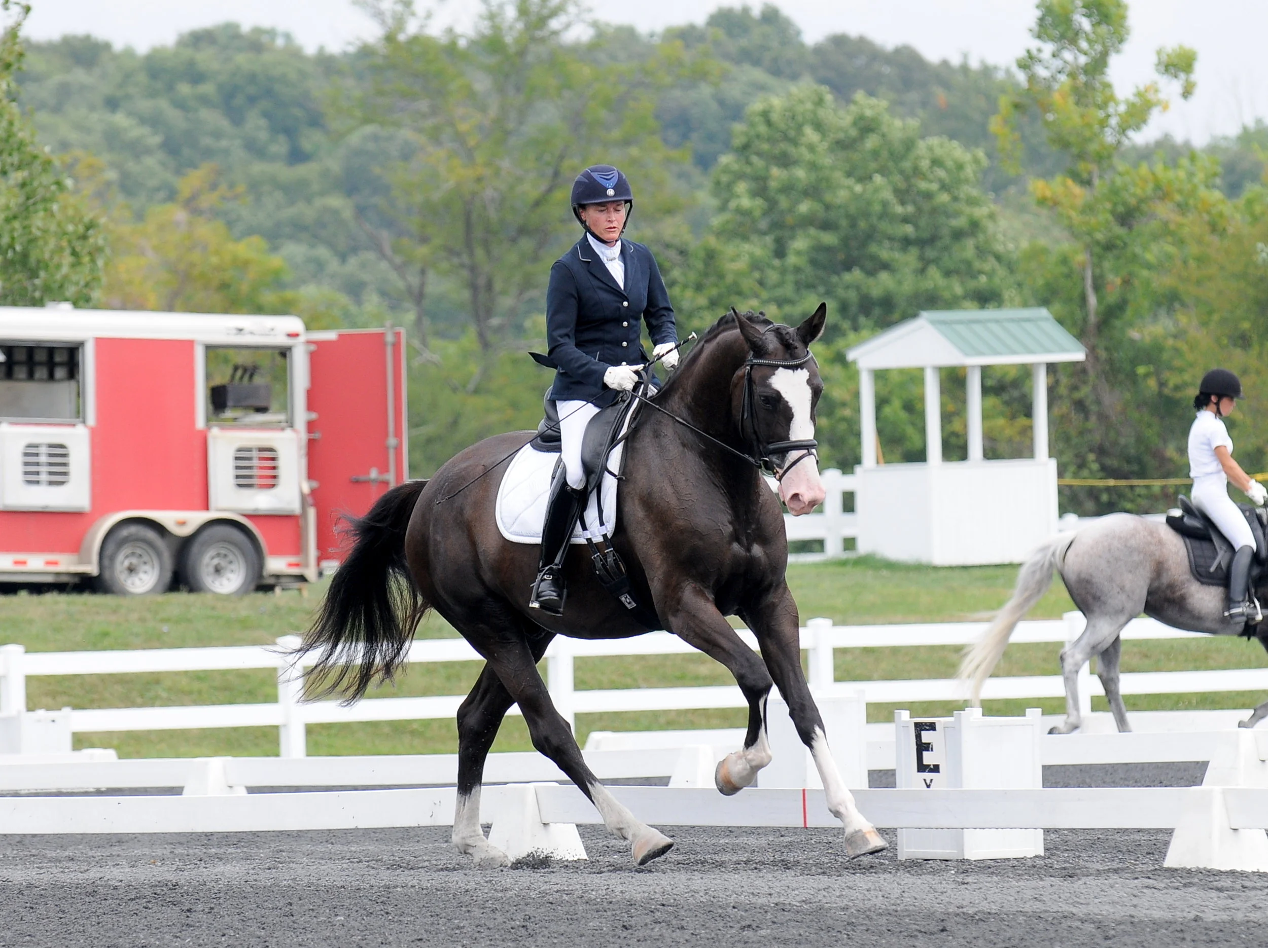 A woman dressed in formal riding attire on a dark horse during a dressage competition, with another rider on a gray horse in the background, set outdoors with trees and a small coach station visible.