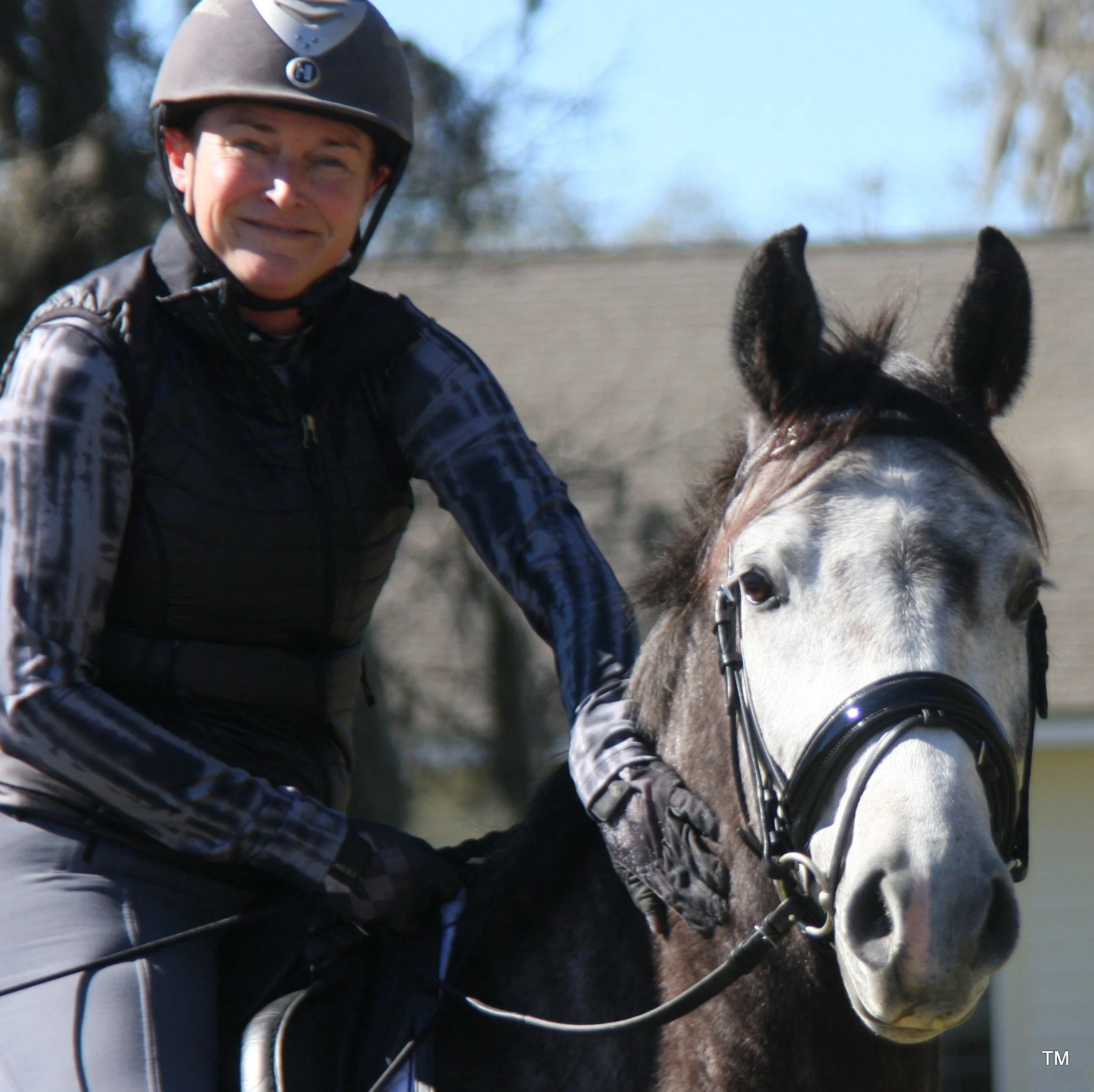 Phoebe DeVoe-Moore wearing a riding helmet and gloves riding a gray and black horse outdoors on a sunny day.