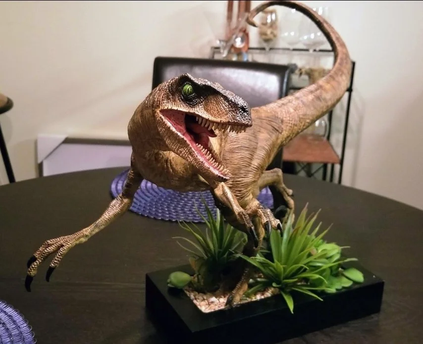 A dinosaur model of a Velociraptor is displayed on a table with green plants at the base, in a room with a chair and household items in the background.