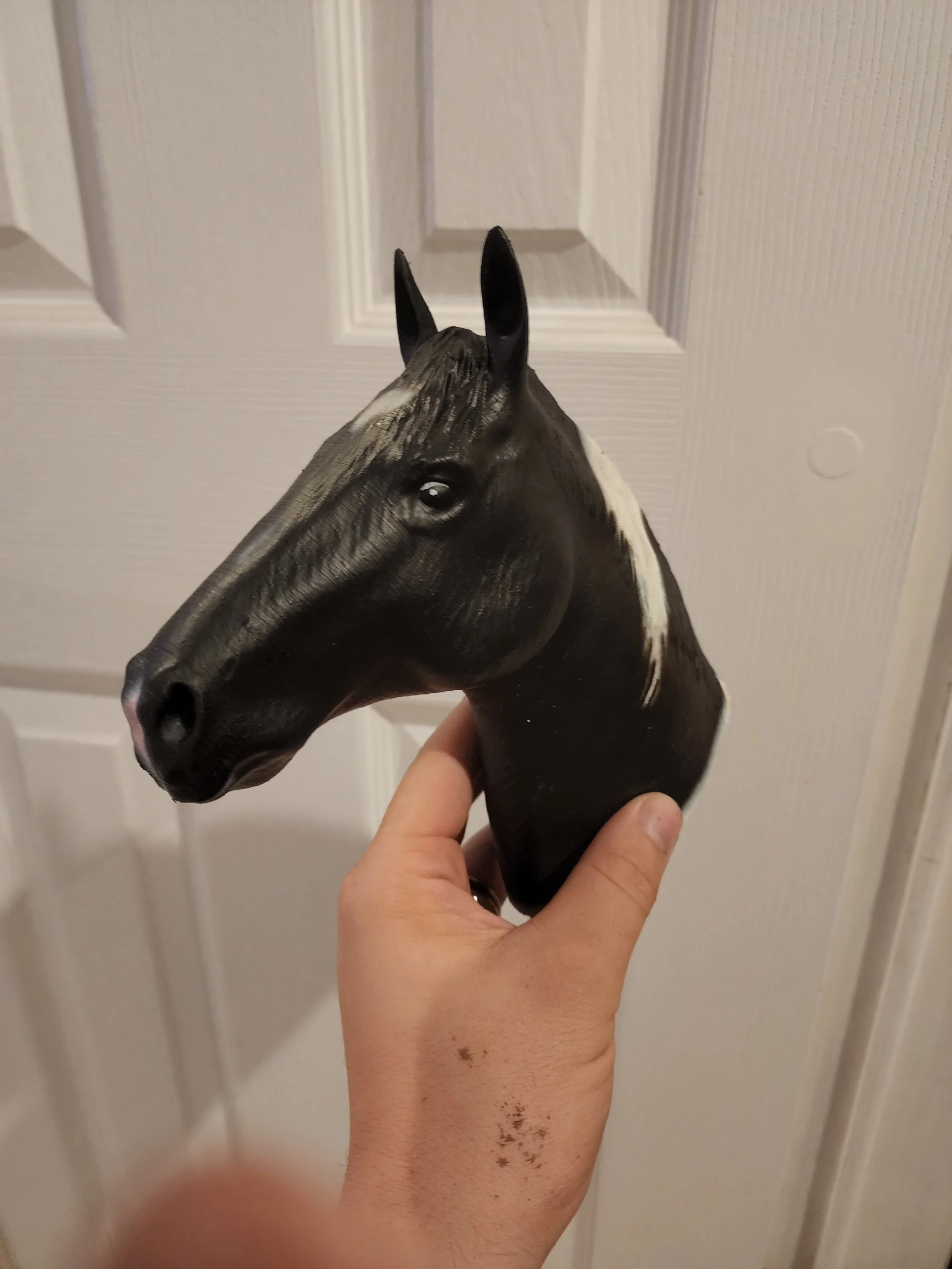 A person holding a horse head sculpture made of a dark material with white markings, standing in front of a white door.