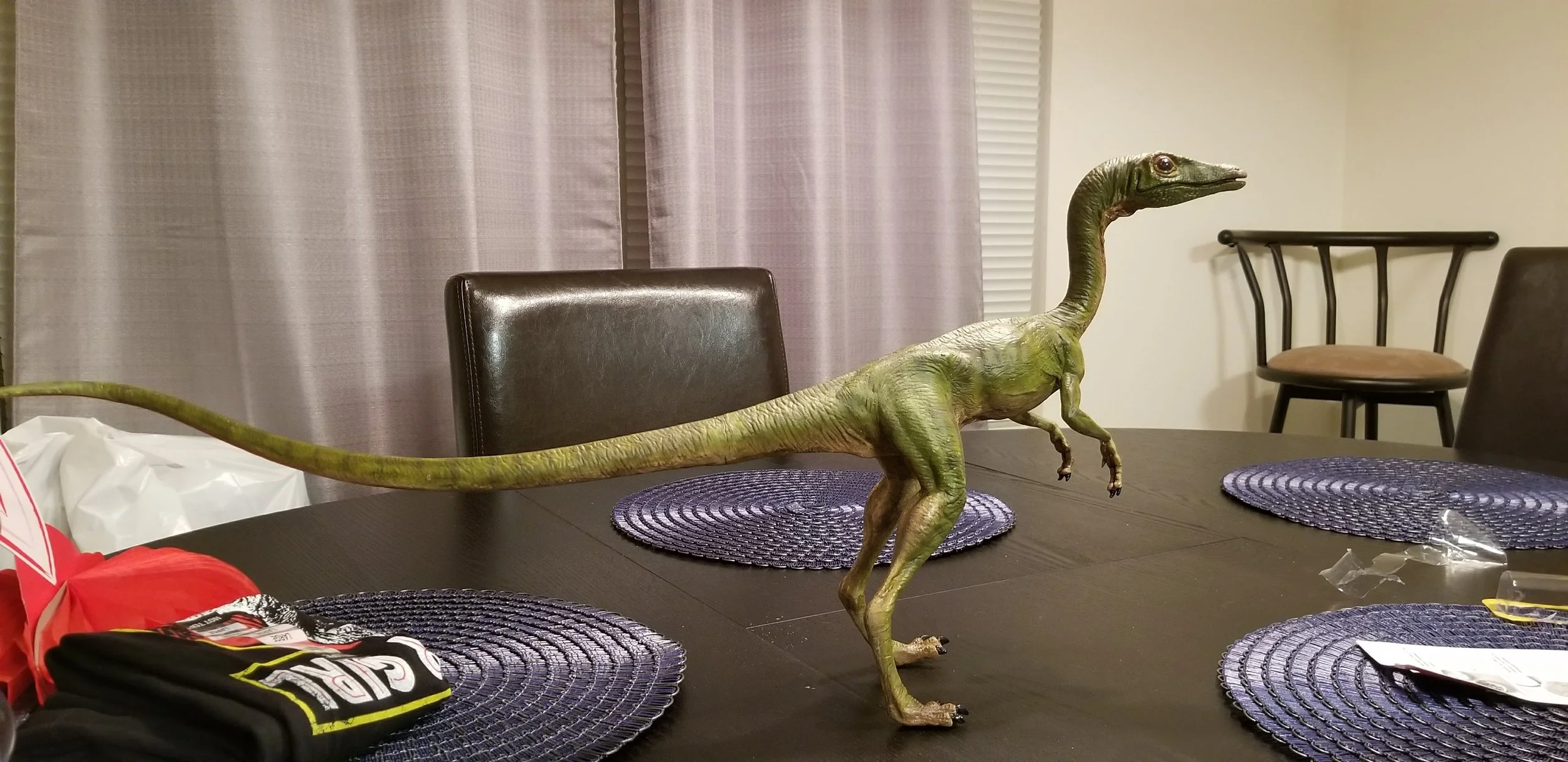 A realistic-looking model Compsognathus, specifically a theropod, stands on a dining table with purple woven placemats. The background includes curtains and chairs around the table.