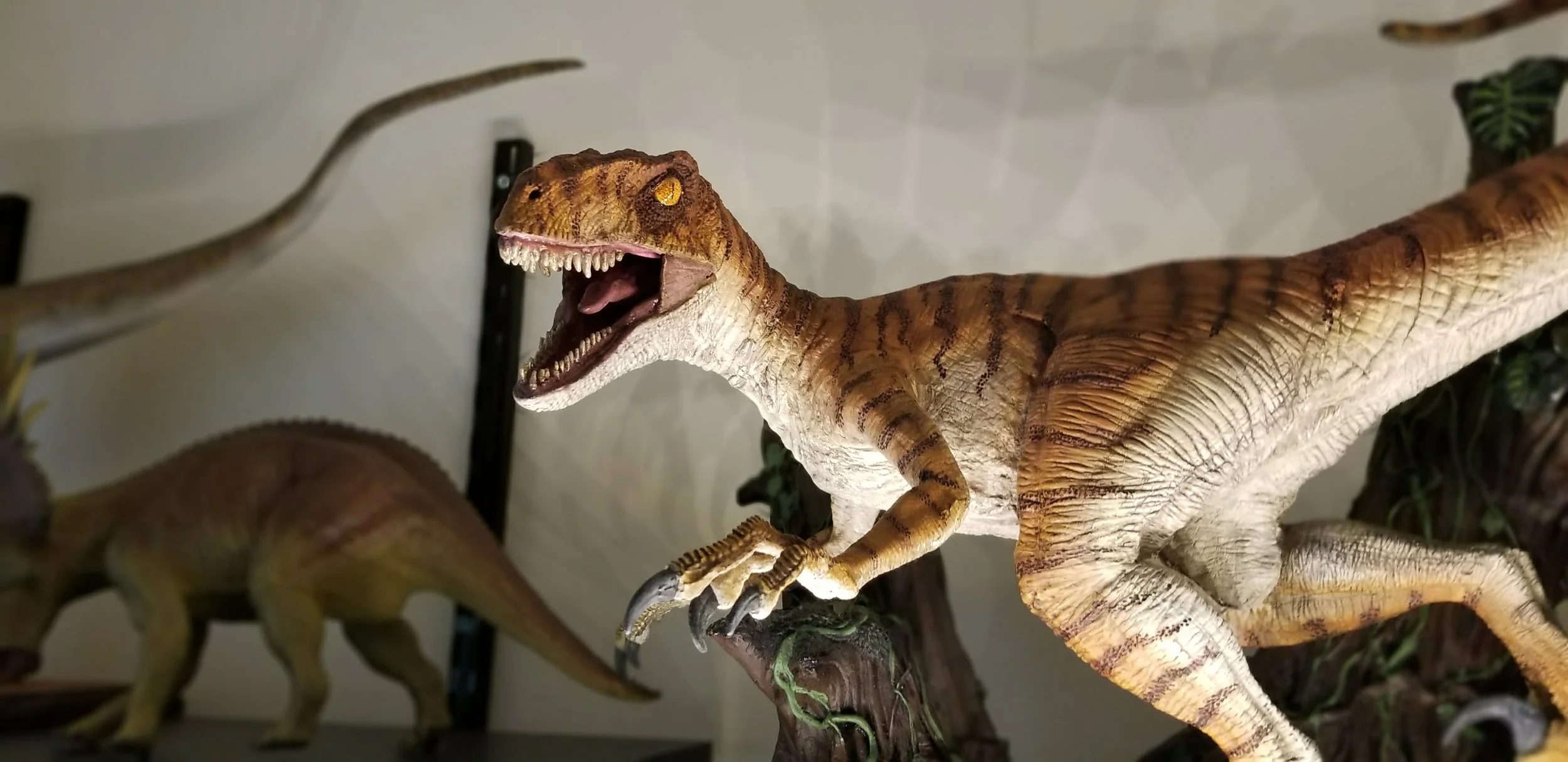 A detailed model of a Velociraptor dinosaur with its mouth open, showing sharp teeth, standing on a branch. Another dinosaur model is partially visible in the background.