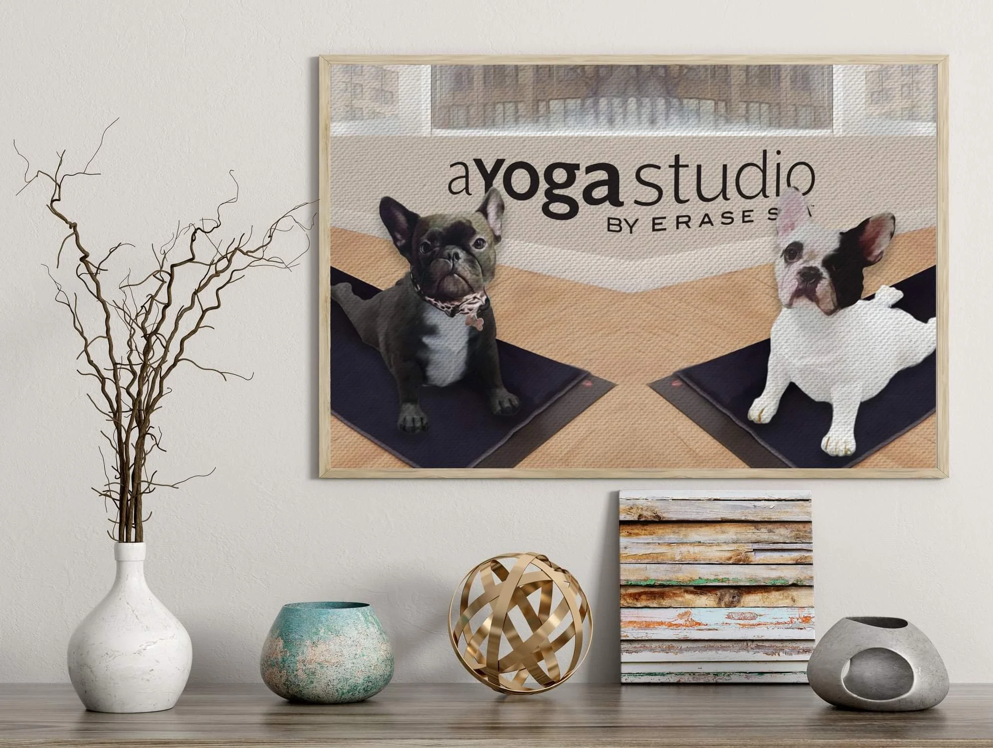 Decorative wall art featuring two French bulldogs on yoga mats, with the text 'a yoga studio by Erases' in the background, hung above a wooden table with decorative objects.