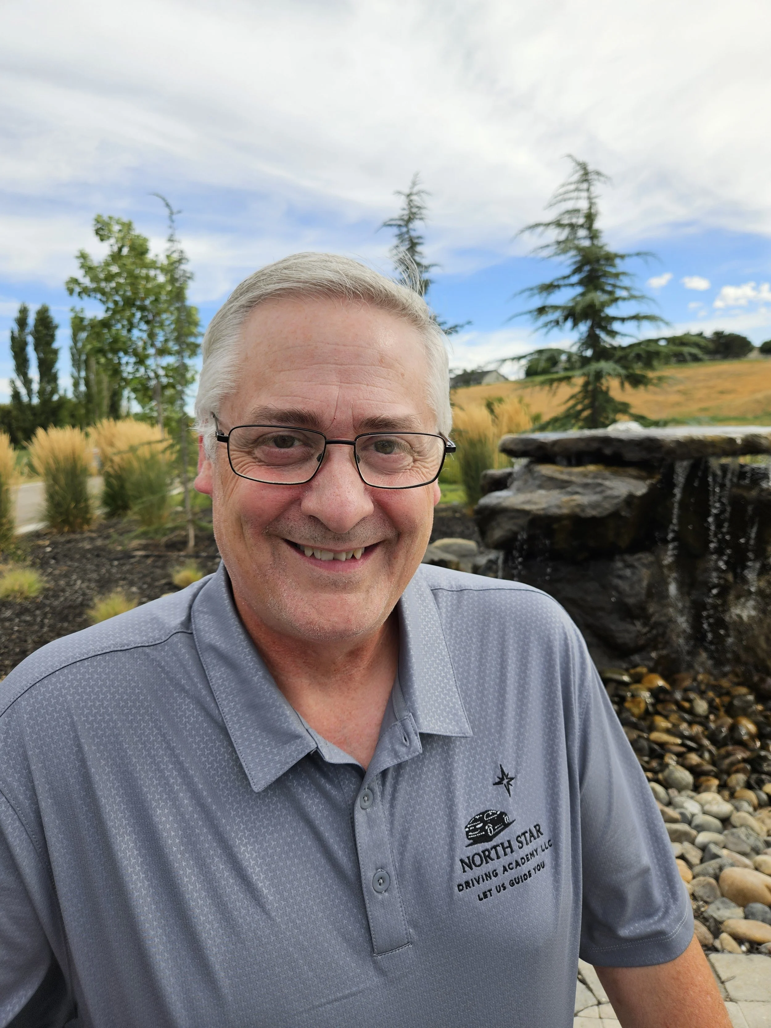 A headshot of driving instructor Nick Schamber in a North Star Driving Academy shirt. He is sitting in front of young trees and a small waterfall installation.