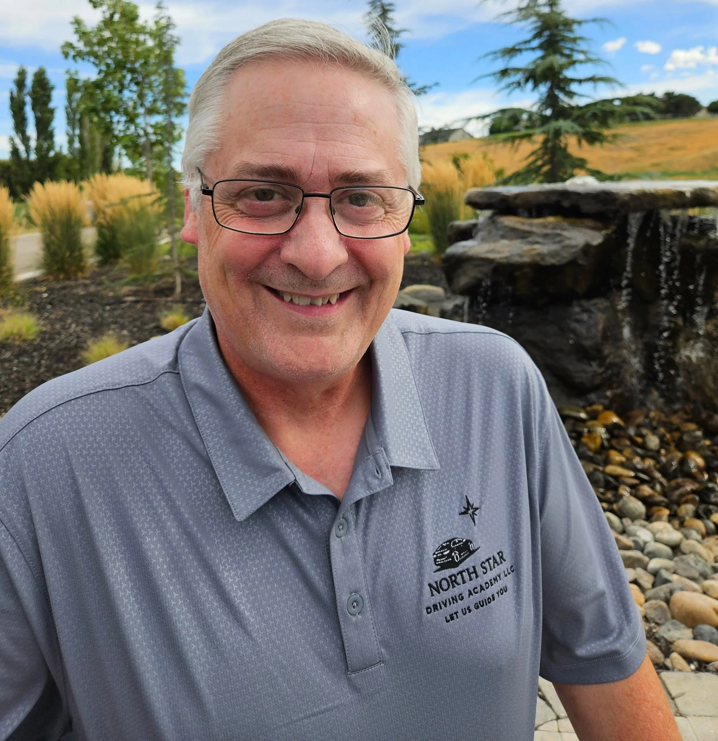 Close-up of a smiling man wearing glasses in an outdoor setting with trees, a sky with clouds, and a rock feature in the background.
