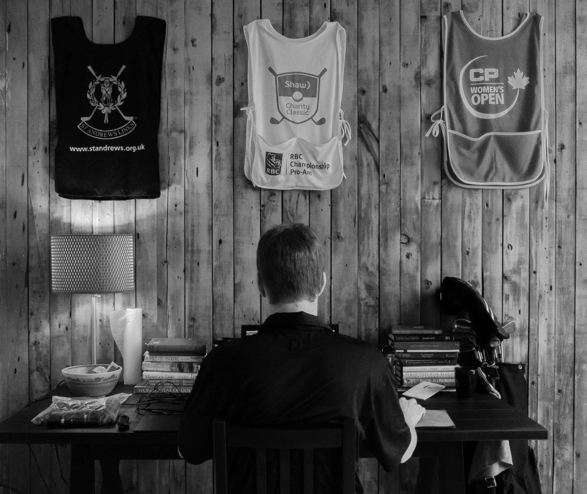 Ben Malach is sitting at a cluttered desk with a stack of books, a golf bag, and a lamp in a room with wooden panel walls.