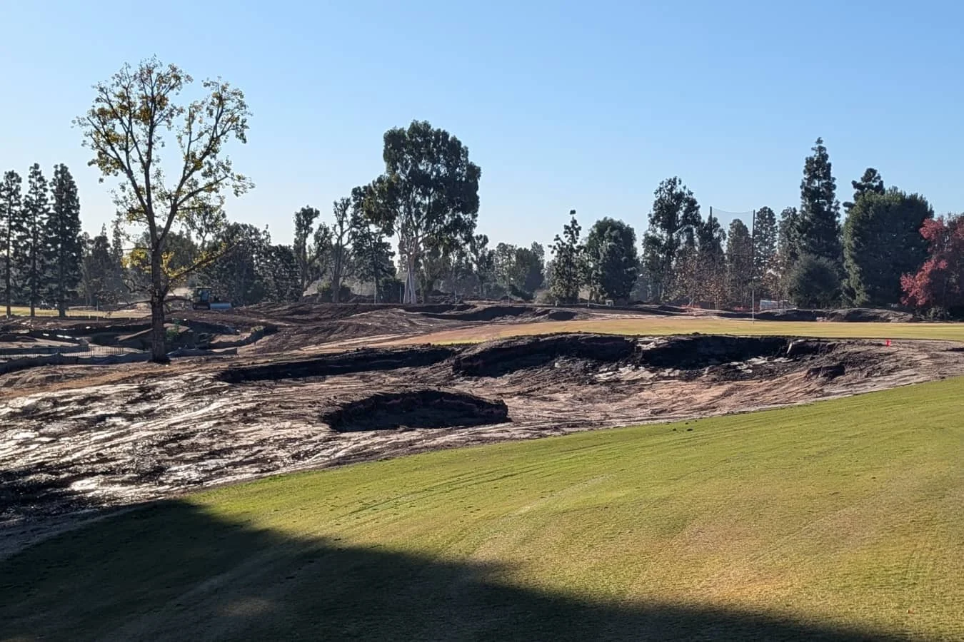 A golf course under construction with disturbed soil, patches of grass, trees in the background, and clear blue sky.