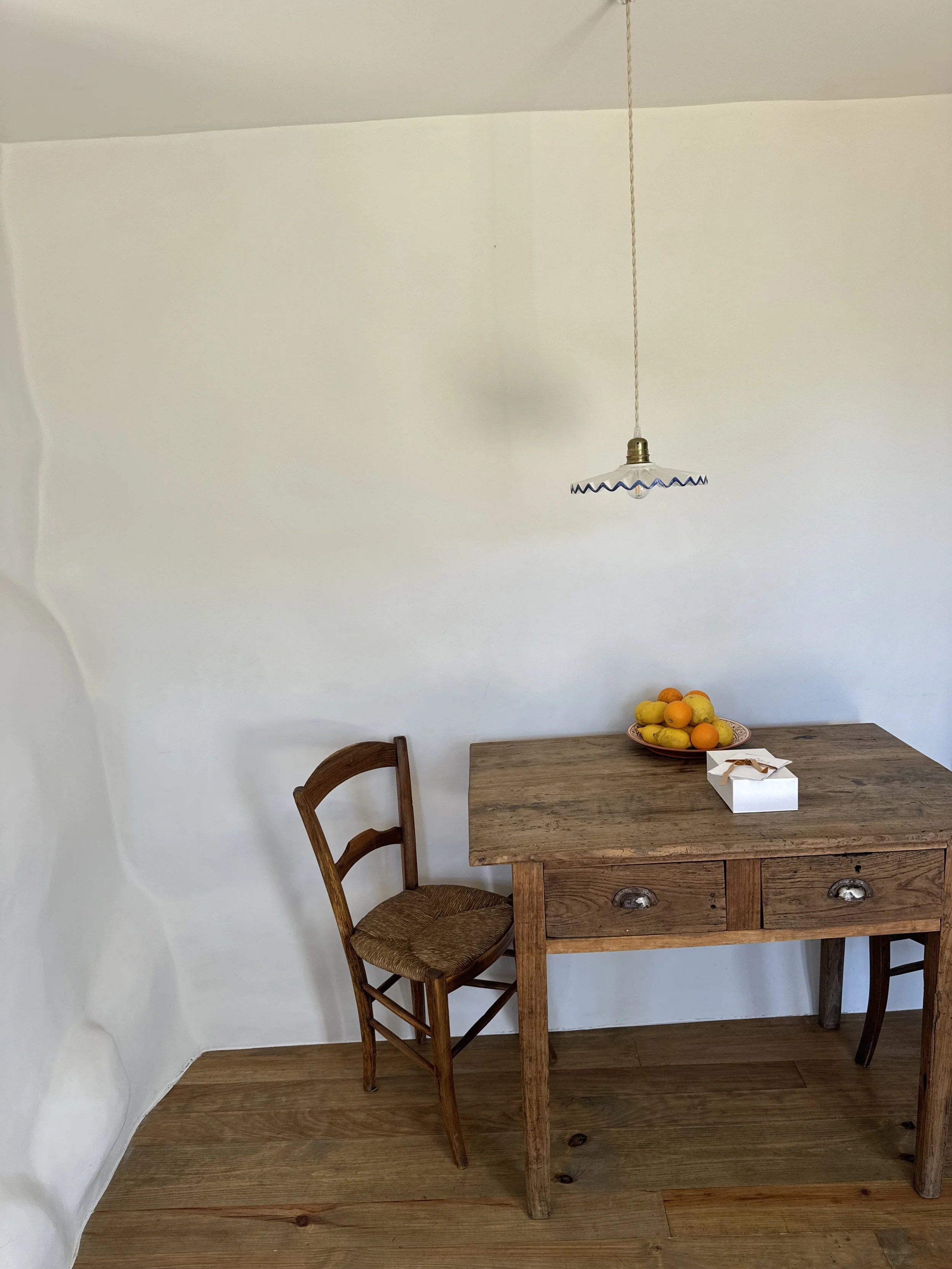 A wooden table with two drawers and a wooden chair with a woven seat. On the table, there is a bowl of assorted fruits and a small white box. A pendant light hangs above the table, and the wall and flooring are made of light-colored wood.
