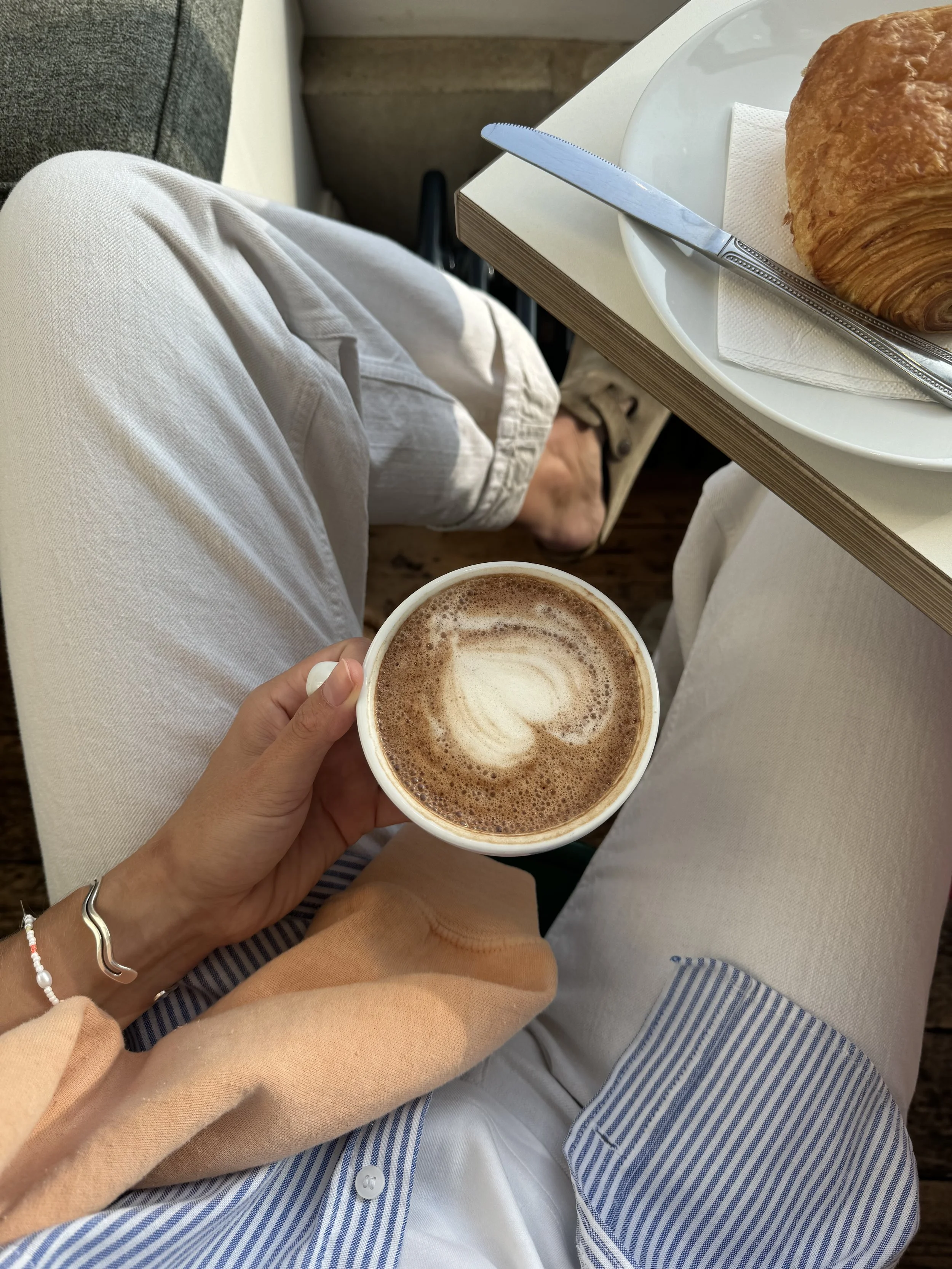 A person holding a cup of coffee with latte art, sitting with legs crossed in a casual setting. On a table nearby, there is a croissant on a white plate with a knife.