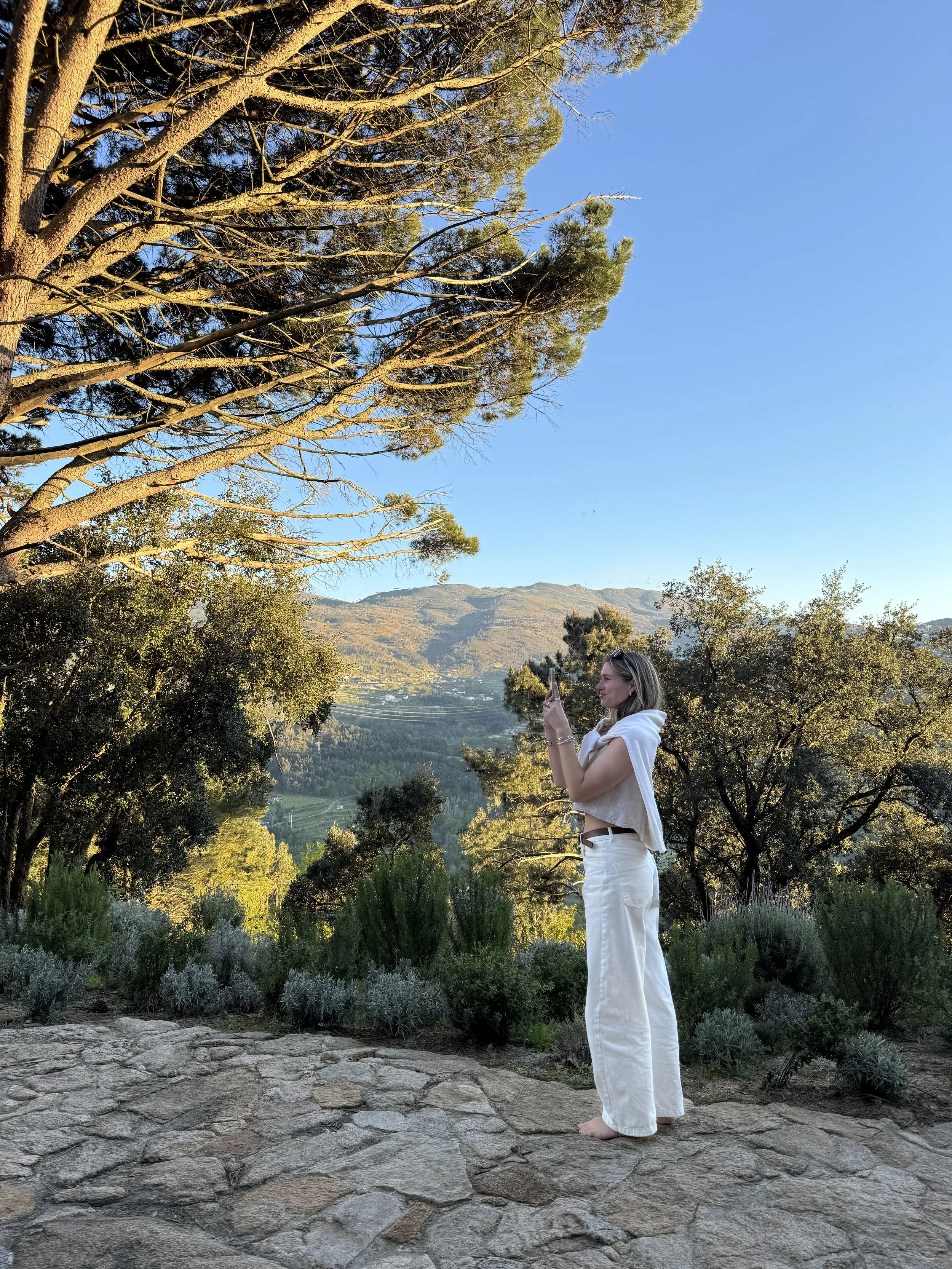 A woman in white pants and a white top, with a covered shoulder, is standing barefoot on a stone path outdoors, taking a photo with her phone. She is surrounded by trees and shrubs, with mountain hills in the background under a clear blue sky.