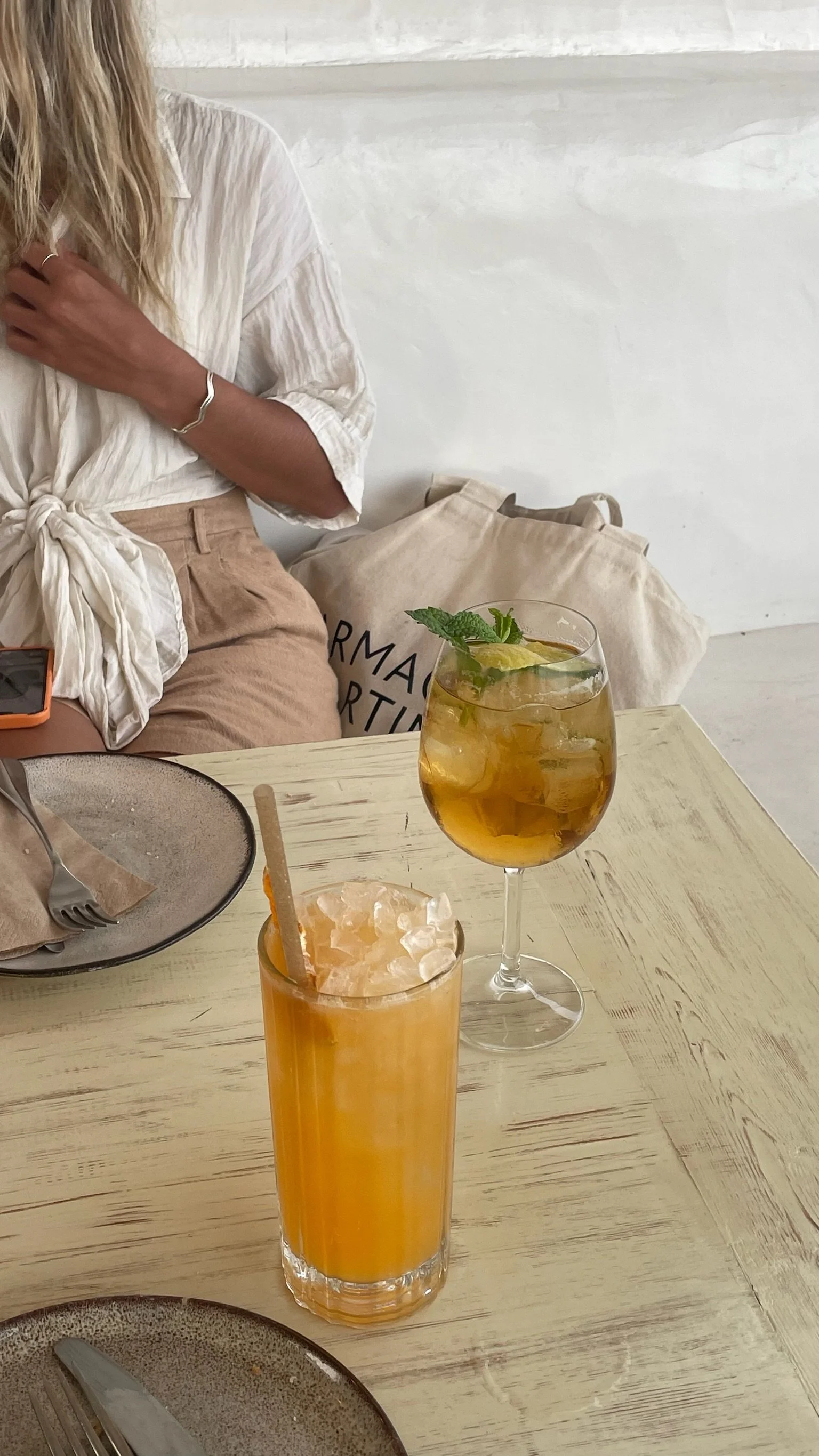 Two beverages on a wooden table; a tall glass of orange juice with ice and a straw, and a wine glass with a yellow cocktail garnished with a lime wedge and mint leaves.