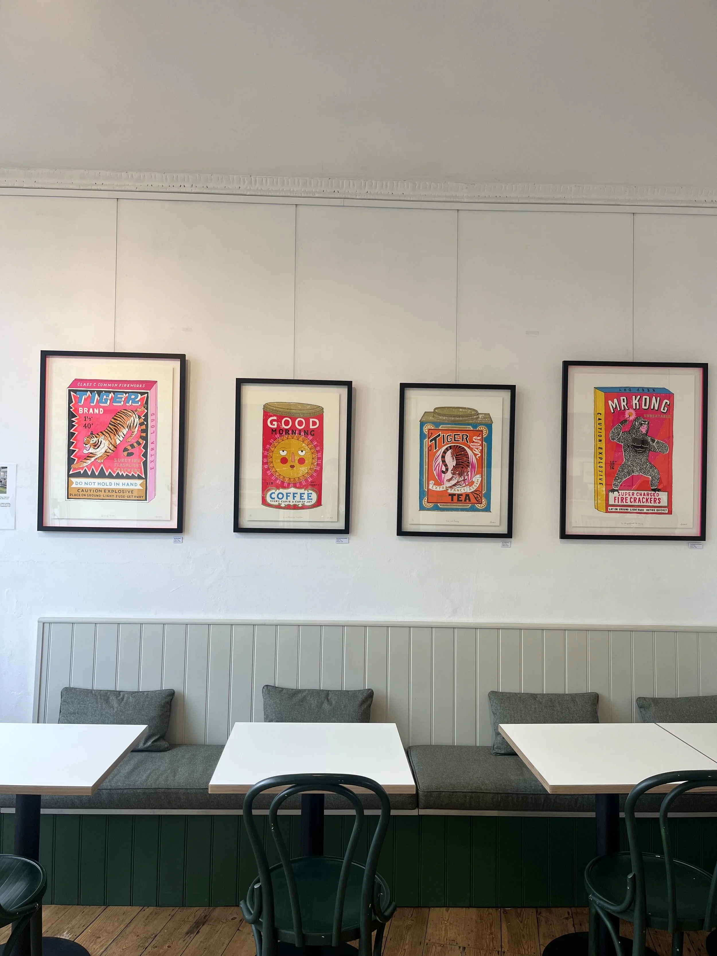 Gallery wall with colorful vintage advertising posters, featuring tiger, coffee, tiger tea, and Mr. Kong, in a cafe with white walls and gray seating.