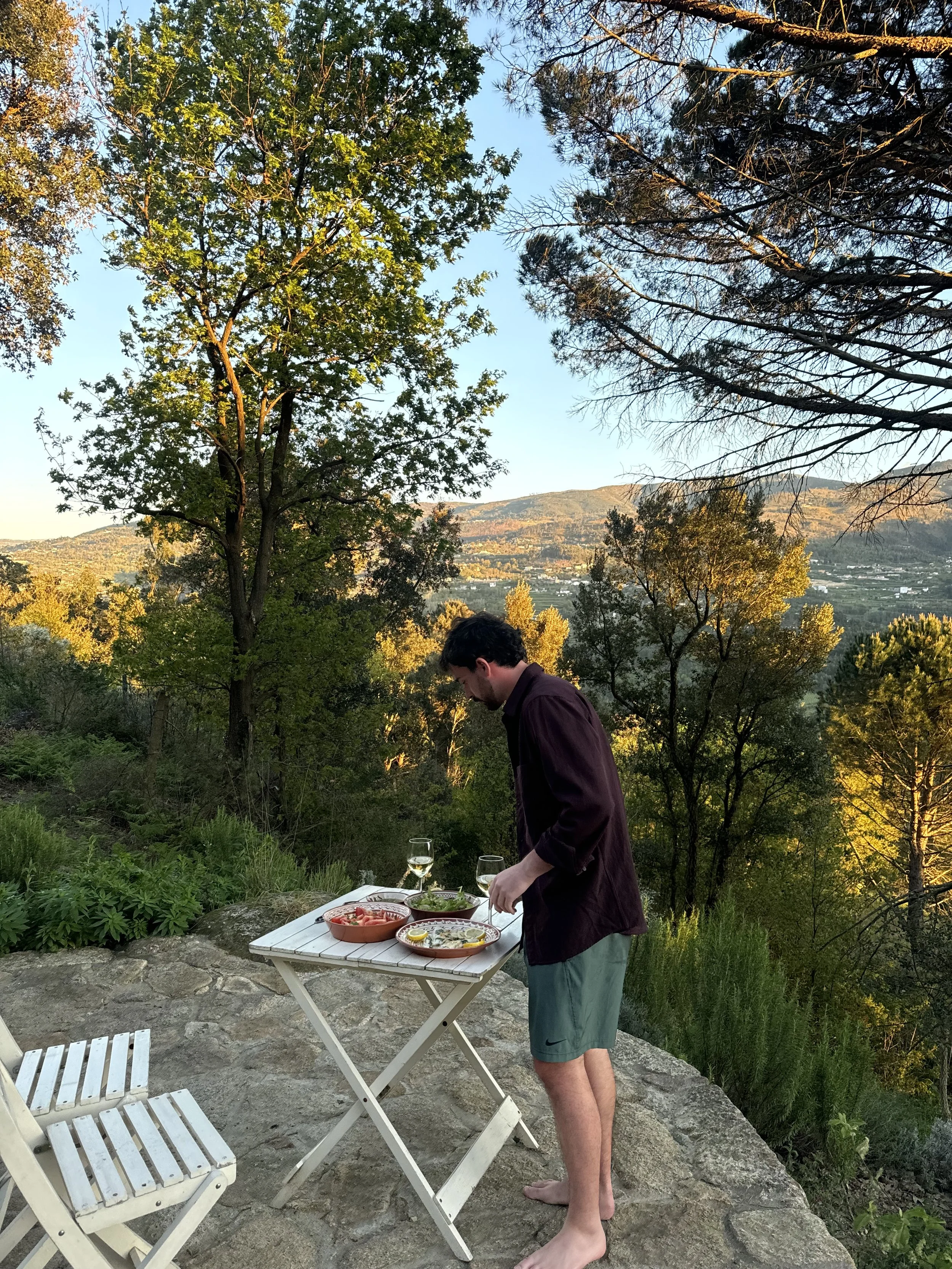 A man preparing food outdoors on a small white table with a scenic landscape of trees and mountains in the background, standing barefoot on a stone surface, with glasses of white wine nearby.