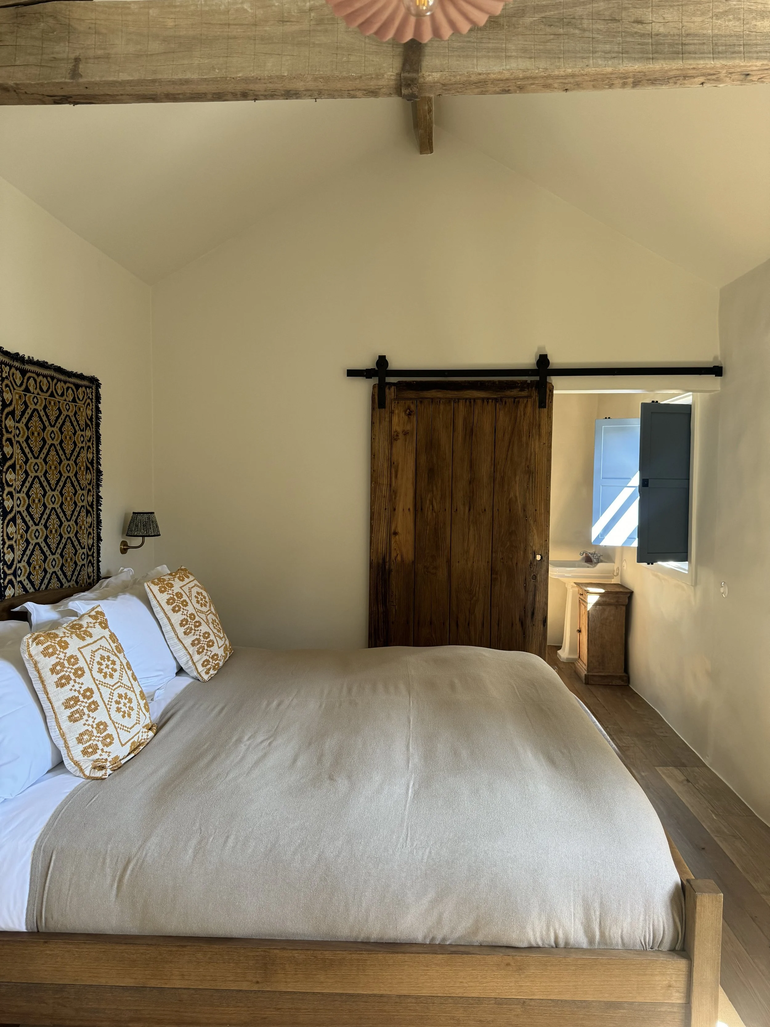 A cozy bedroom with a wooden bed frame and beige bedding, decorated with embroidered pillows. There is a small bedside table next to a window with open blue shutters, and a rustic wooden sliding door.