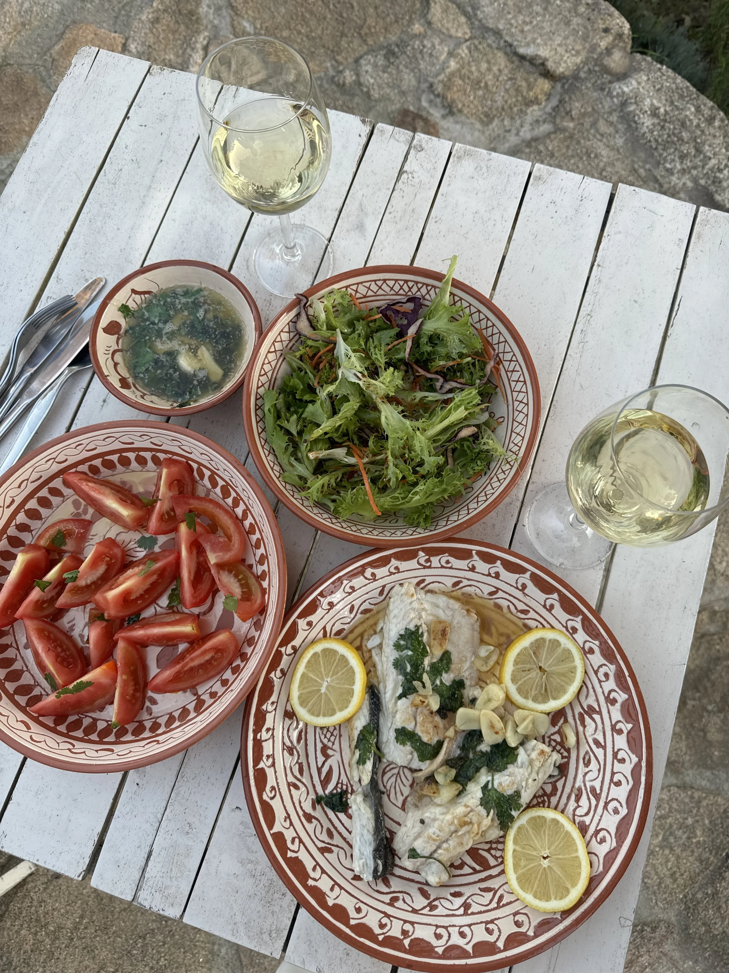 A table set with a meal including a plate of grilled fish garnished with lemon slices and herbs, a bowl of mixed green salad, a bowl of tomato wedges, a small bowl of soup, and two glasses of white wine on a white wooden table outdoors.