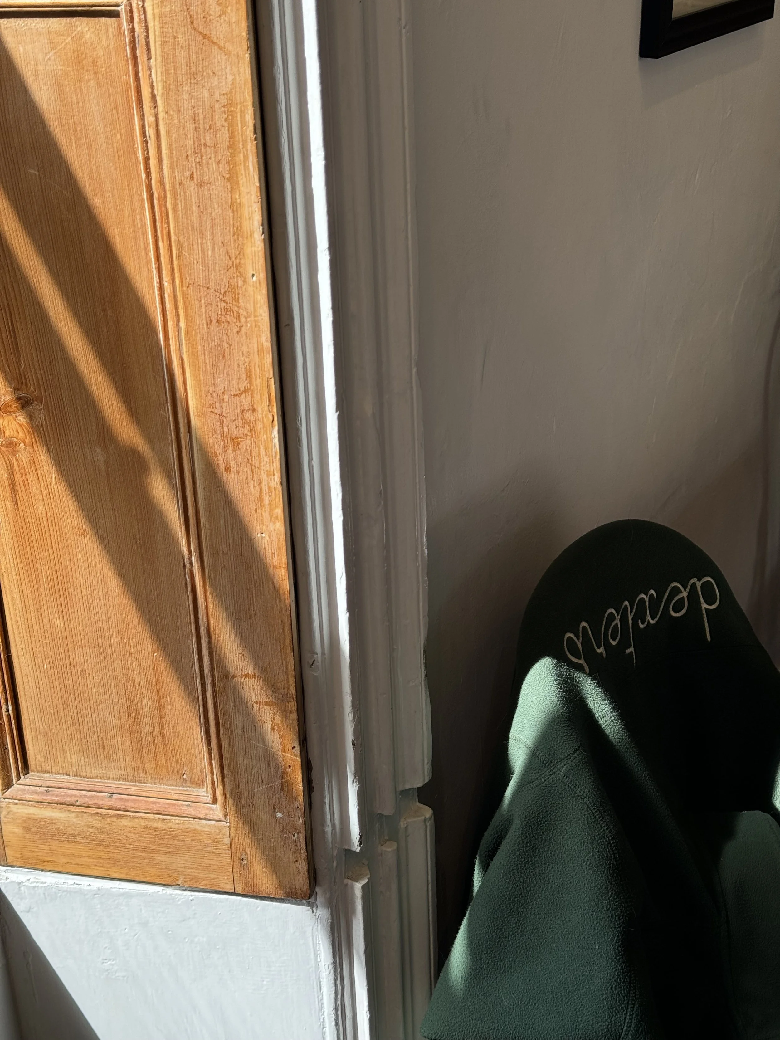 Close-up of a doorframe and baseboard with sunlight casting shadows, a person wearing black pants with a cursive written word, and a green bag or purse resting against the wall.