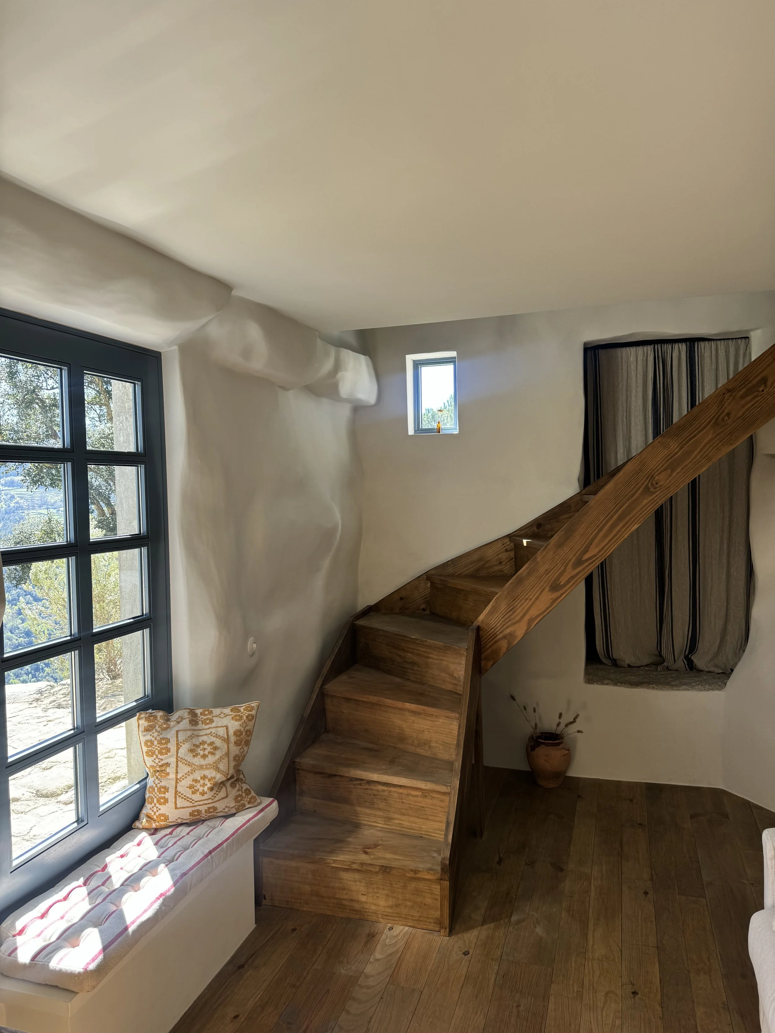 Interior of a cozy house with white walls, wooden staircase, window seat with cushion and pillow, small side window, small curtain, and wooden flooring.
