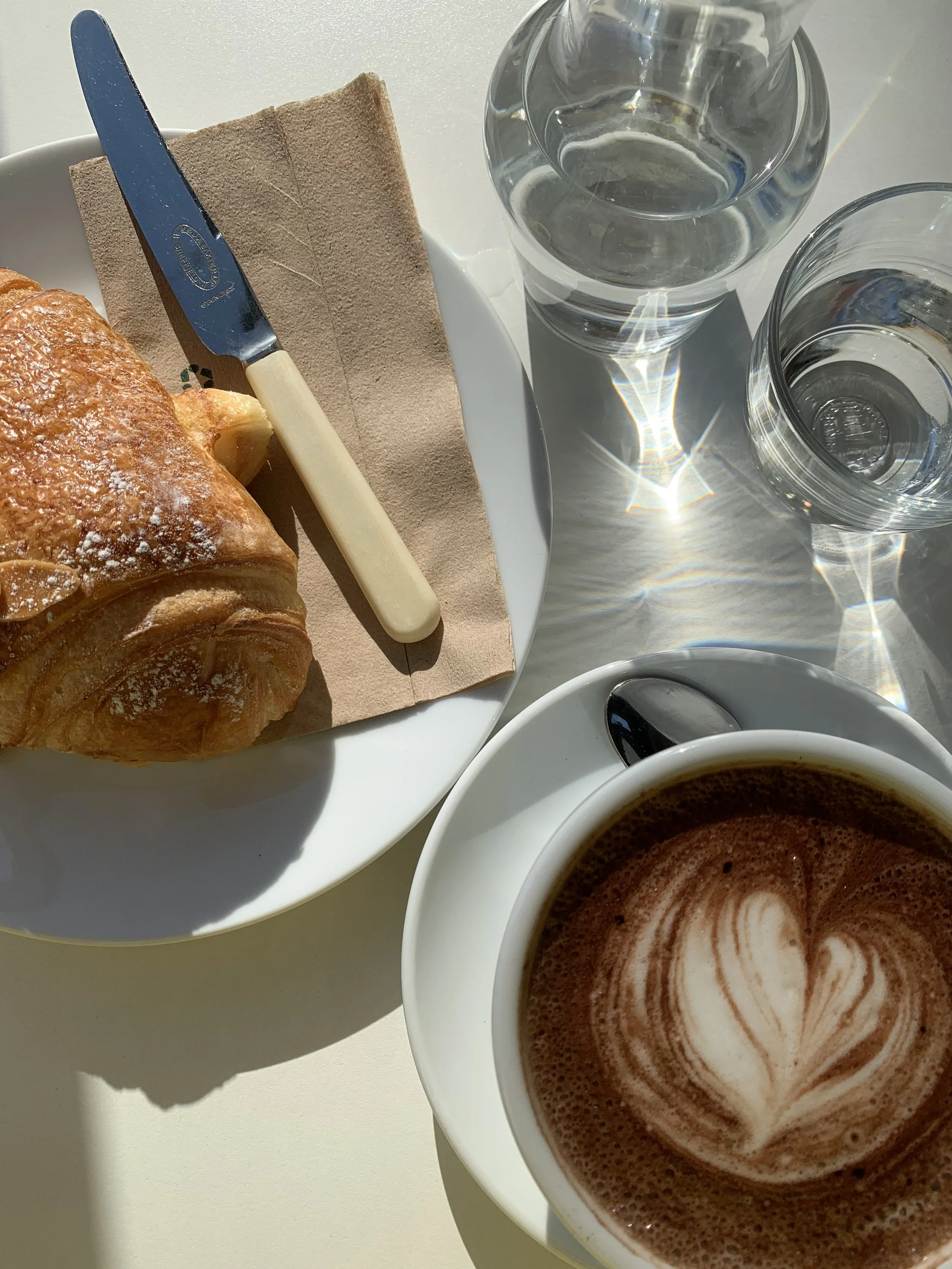 A breakfast scene with a croissant on a white plate, a butter knife with a beige handle, two glasses of water, a cup of coffee with latte art, and a spoon, on a white surface with sunlight and shadows.
