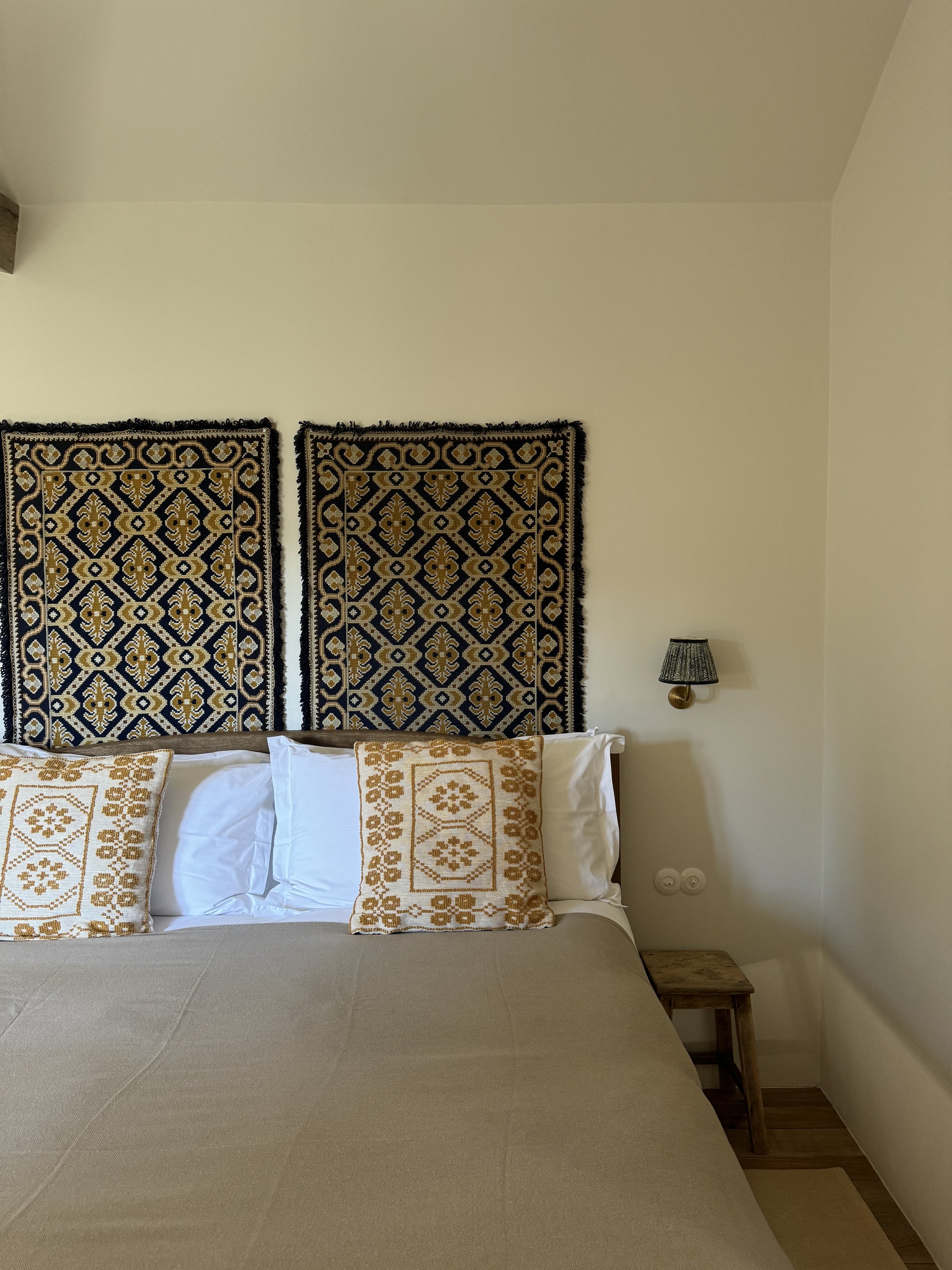 A neatly made bed with beige bedding, two decorative pillows with gold embroidery, a wooden step stool beside the bed, and a wall-mounted lamp to the right. Two patterned textile wall hangings with black, gold, and cream colors are above the bed on a