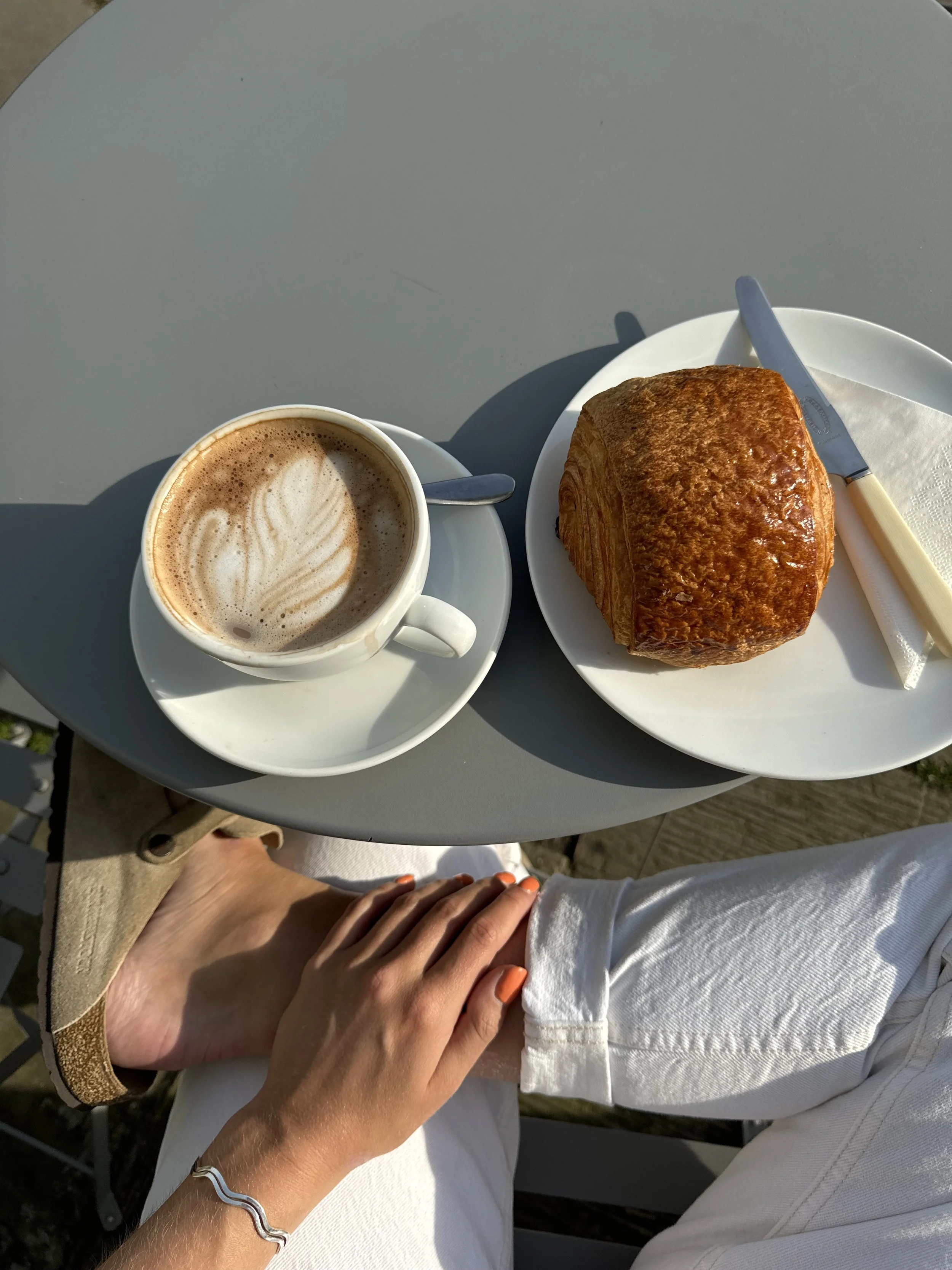 Top-down shot of a latte with latte art, a croissant on a plate, and a person's hand resting on their knee, with a gray table and wooden deck in the background.