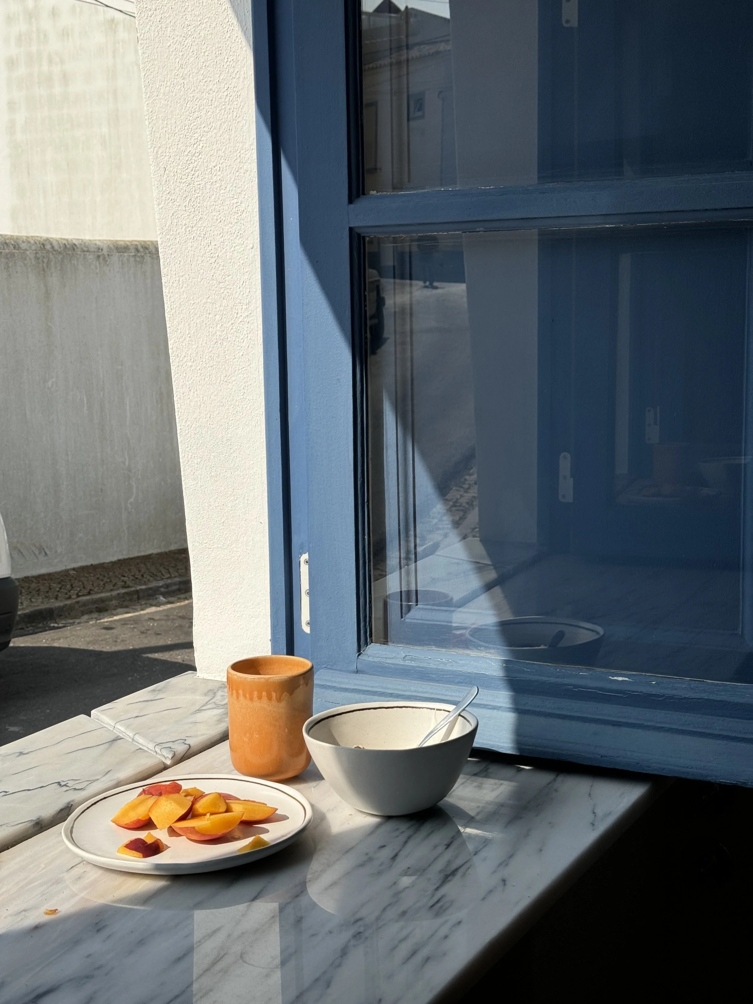 A marble table next to a blue window with a bowl, a ceramic mug, and a plate of sliced peaches and plums, with sunlight casting shadows.