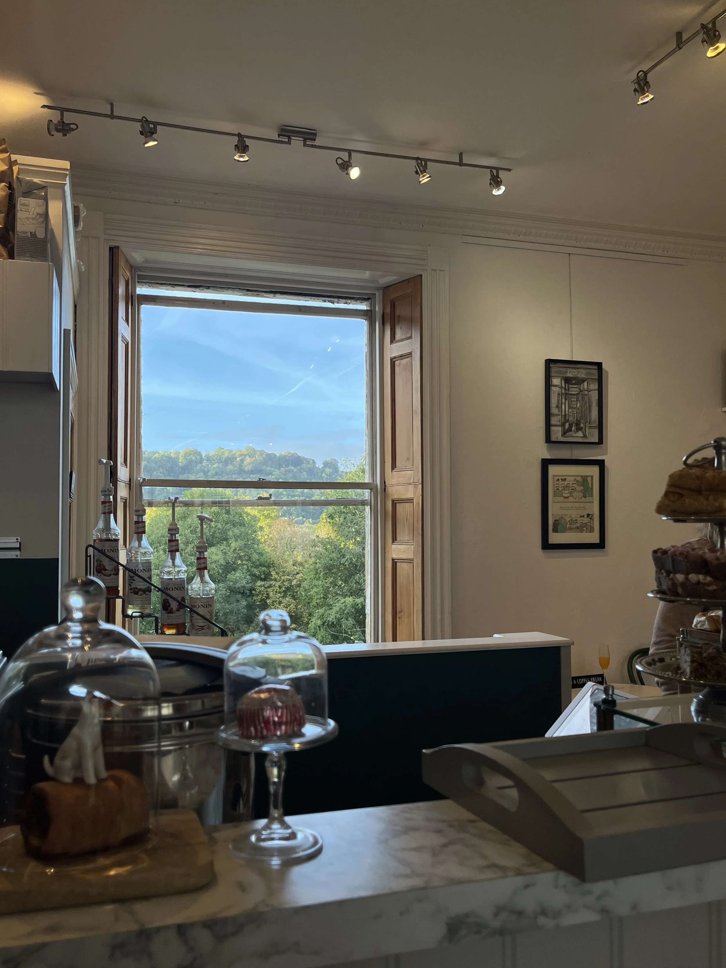 Interior of a cozy cafe with large window showing green trees and blue sky outside. The counter has various baked goods under glass domes, and bottles of alcohol are visible behind the counter. The decor includes framed pictures on the wall and track