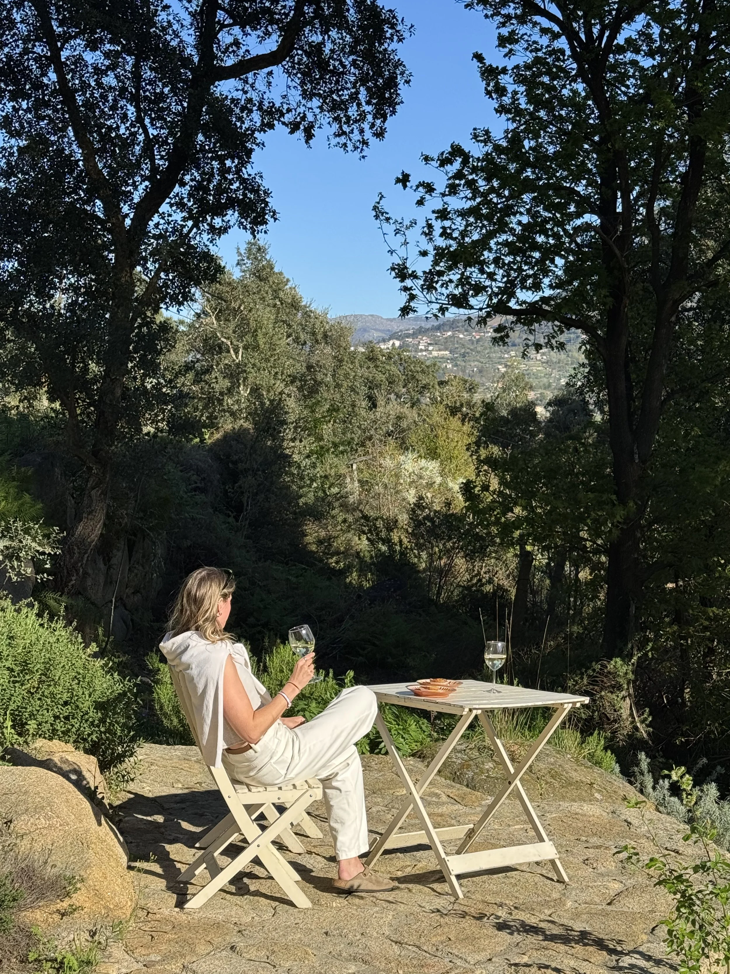 A woman sitting outdoors on a white folding chair, holding a glass of white wine, at a small white table with another glass of white wine and some snacks, surrounded by trees and overlooking a hilly landscape.