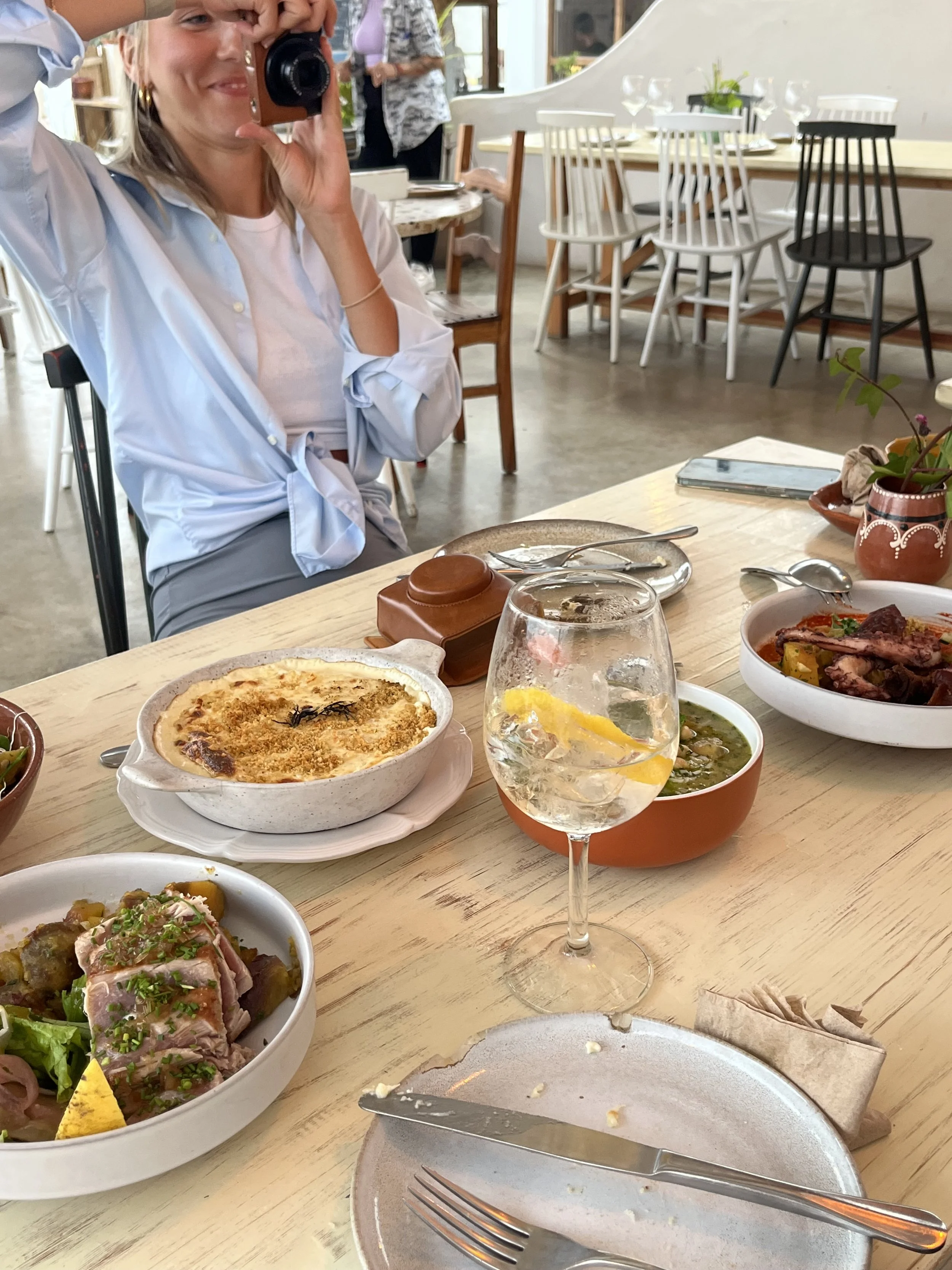 A woman taking a photo of a dining table full of food, including a baked dish, a bowl of salad, a bowl of soup, and some other plates, in a bright, modern restaurant with other tables and chairs visible in the background.