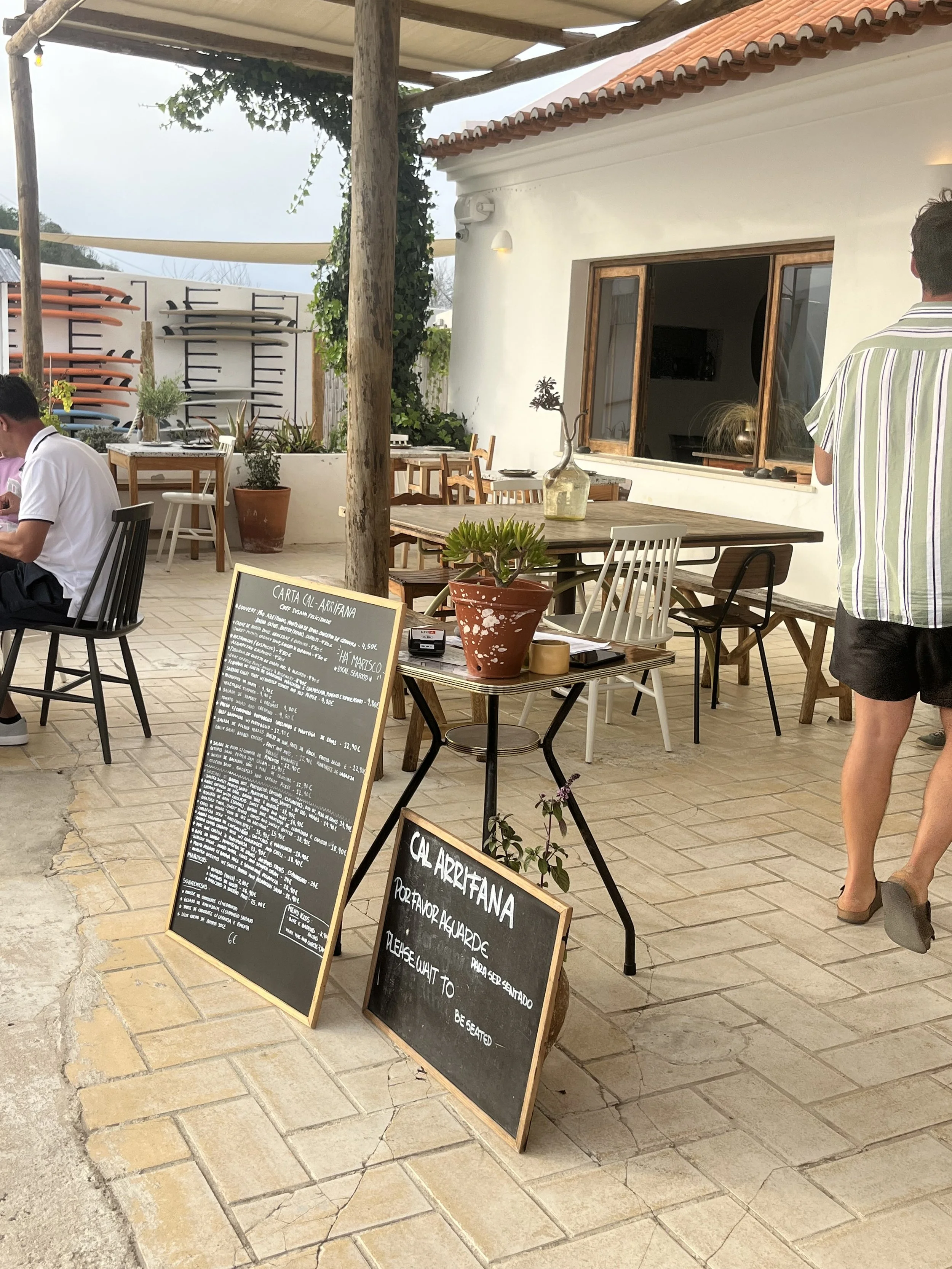 Outdoor restaurant with tables, chairs, potted plants, menu boards, and a white building with an open window.