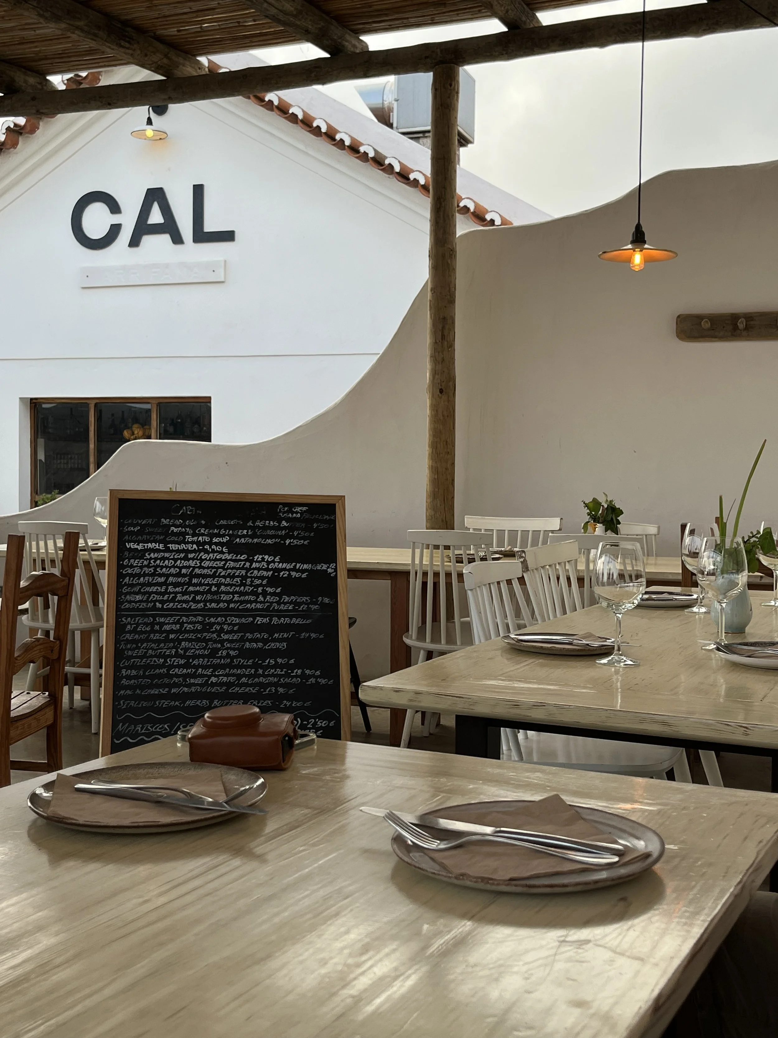 Interior of a restaurant with wooden tables and white chairs, a chalkboard menu, and a white building with the letters CAL on the wall outside.
