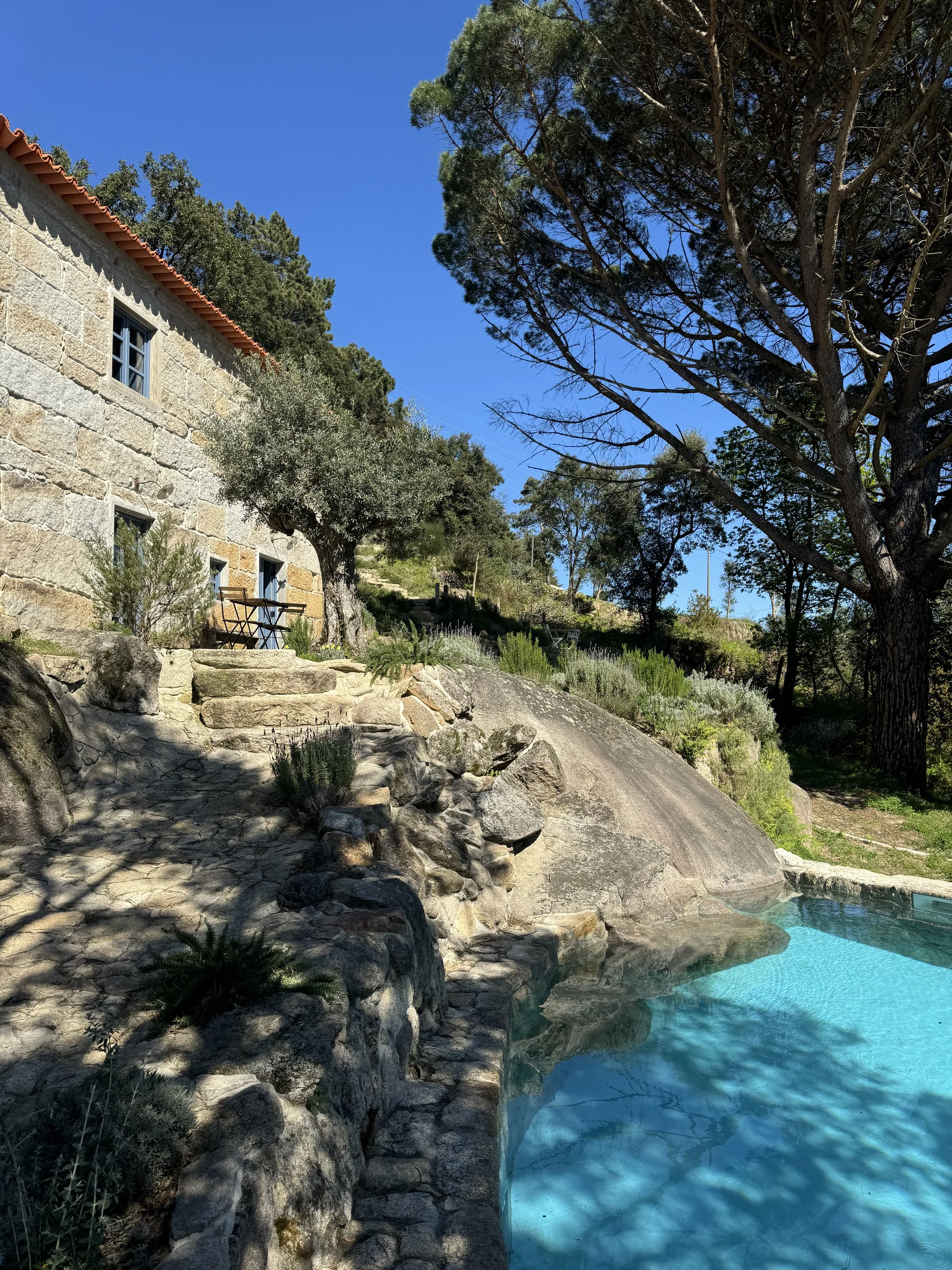 A stone house next to a rocky landscape with a swimming pool, trees, and clear blue sky.