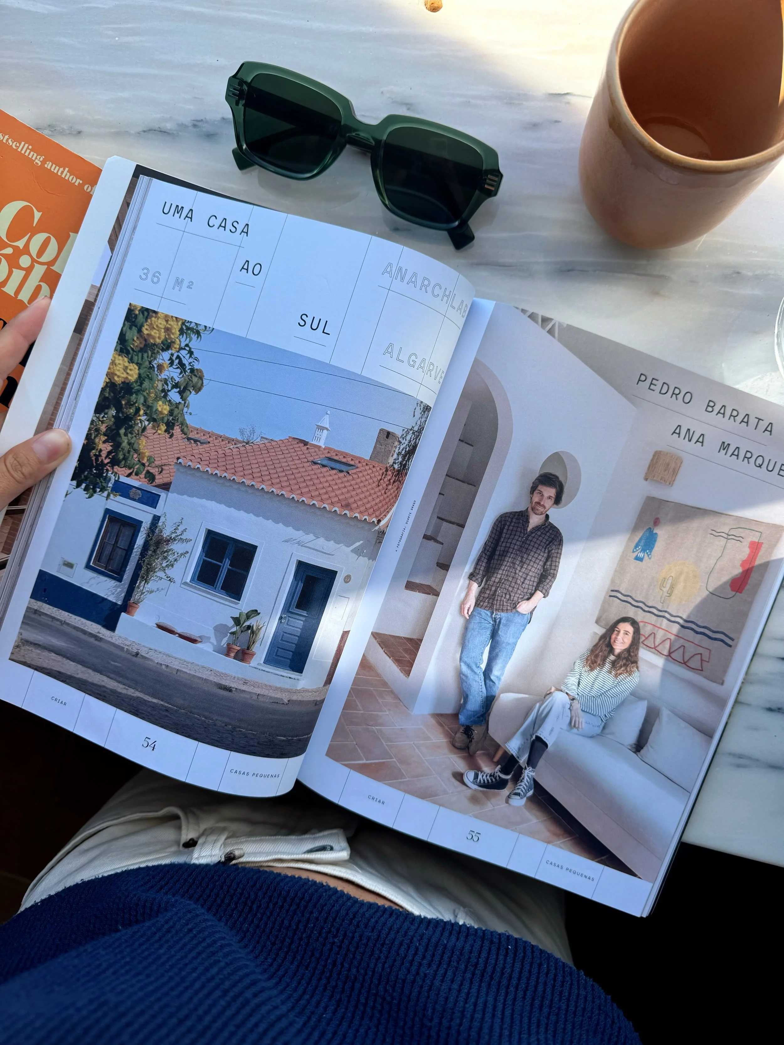 Open magazine showing a photo of a white house with a red tile roof, green plants, and blue window frames, and a photo of two people sitting and standing inside a room with a colorful wall hanging.