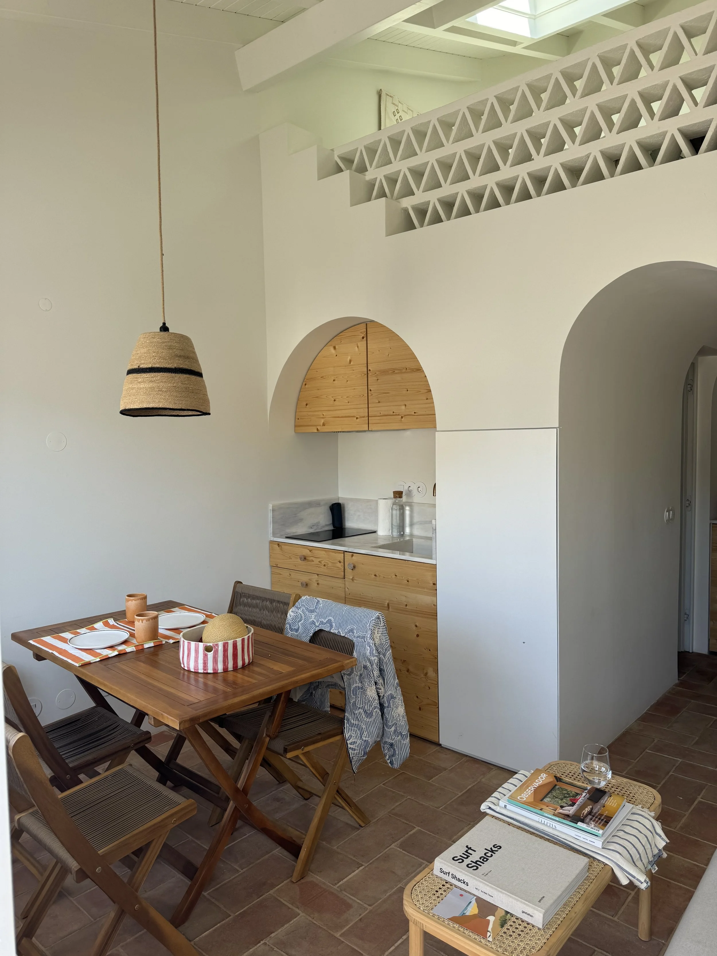 Dining area with a wooden table set with plates and cups, surrounded by chairs, near a small kitchen with wooden cabinets, and a corner table with magazines and a glass of water.