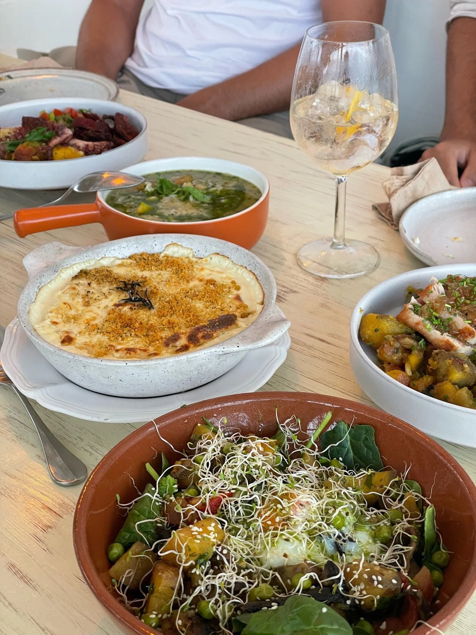 Assorted dishes of Mexican cuisine on a light wood table, including a salad with sprouts, a baked cheesy dish, a bowl of green soup, and other bowls with mixed vegetables and meat, with a person in a white shirt in the background.
