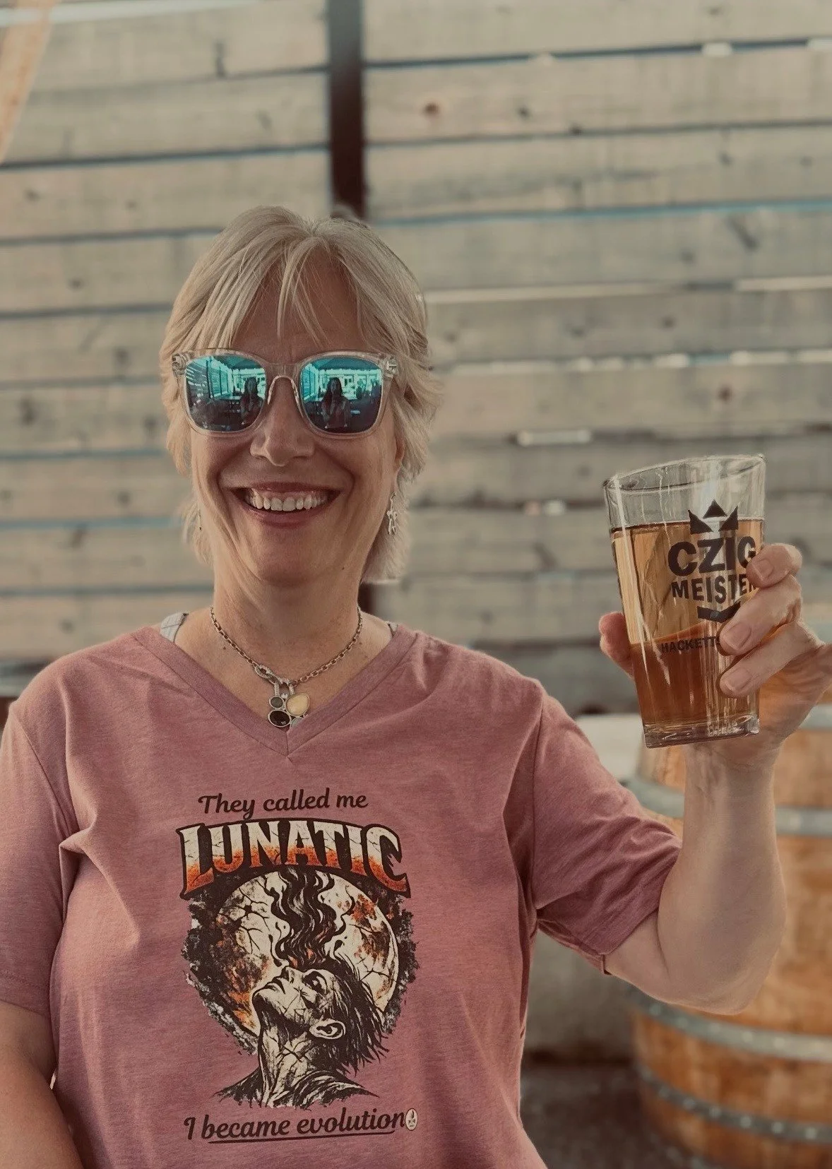 A smiling woman with short blonde hair and sunglasses holding a glass of beer in a brewery with wooden walls in the background.