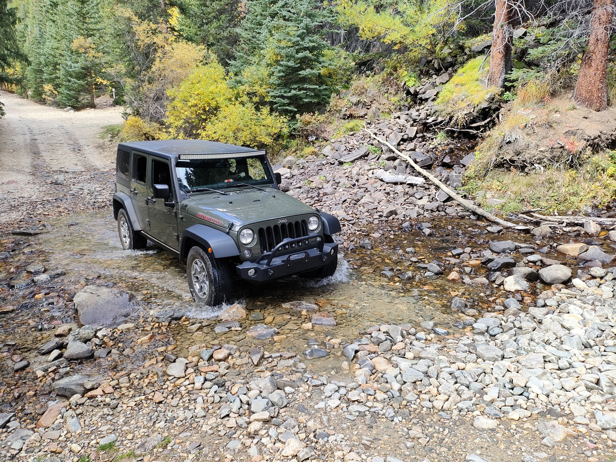 Green 2015 Jeep Wrangler Unlimited Rubicon rental driving through a shallow rocky creek in a forested area in Colorado
