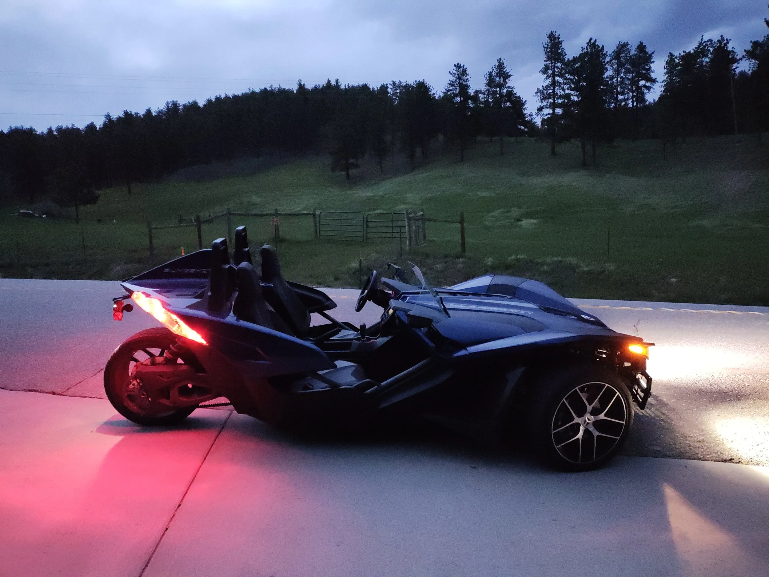 Blue 2018 Polaris Slingshot roadster with three seats, parked on the side of a road at dusk, with a grassy hill and trees in the background in Highlands Ranch, Colorado