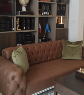 A brown tufted sofa with two green pillows in a room with a bookshelf and a small table with a chessboard.