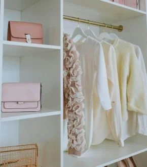 Closet with hanging white and cream clothes, pink handbags on white shelves, and a gold clothing rod.